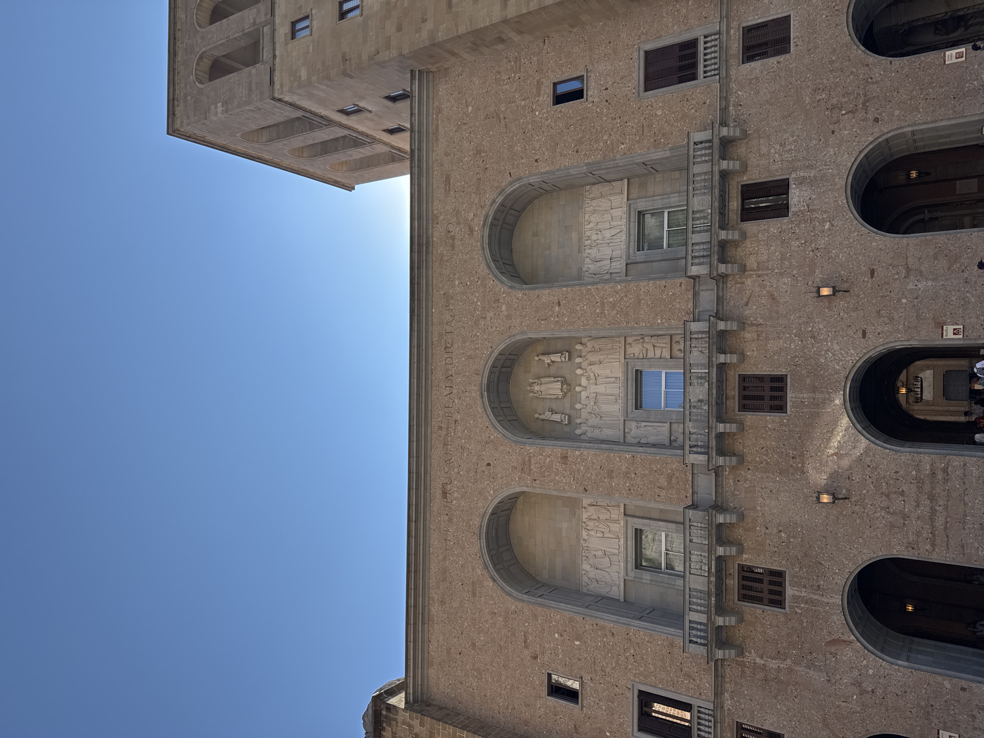 The photograph captures a low-angle view of the facade of the Montserrat Abbey in Monistrol de Montserrat, Spain, under a clear blue sky. The building is constructed from light brown stone with a speckled texture. The main section of the building features a row of three large arched alcoves, each adorned with relief sculptures. The central alcove contains a sculpture of figures standing on a pedestal, while the flanking alcoves depict rows of standing figures. Above these alcoves, an inscription is carved into the stone, part of which is visible as "I.E.S. JERUSALEM BEATA D..." and then another section reads "PATRONA. SUA. CRISTIANA. O. NOSTRA. VIRGINIS. SANCTAE".
Below the alcoves, a balcony with stone balusters runs across the facade, supported by decorative brackets. There are several windows with dark shutters on this level and on the upper stories. To the right, a taller, tower-like section of the abbey rises, also constructed of stone and featuring multiple arched openings. The lower part of the facade shows several large arched entrances leading into the building, with lampposts visible in the shadows. A few people are partially visible entering or standing near these lower arches. The sunlight casts subtle shadows on the building's features.