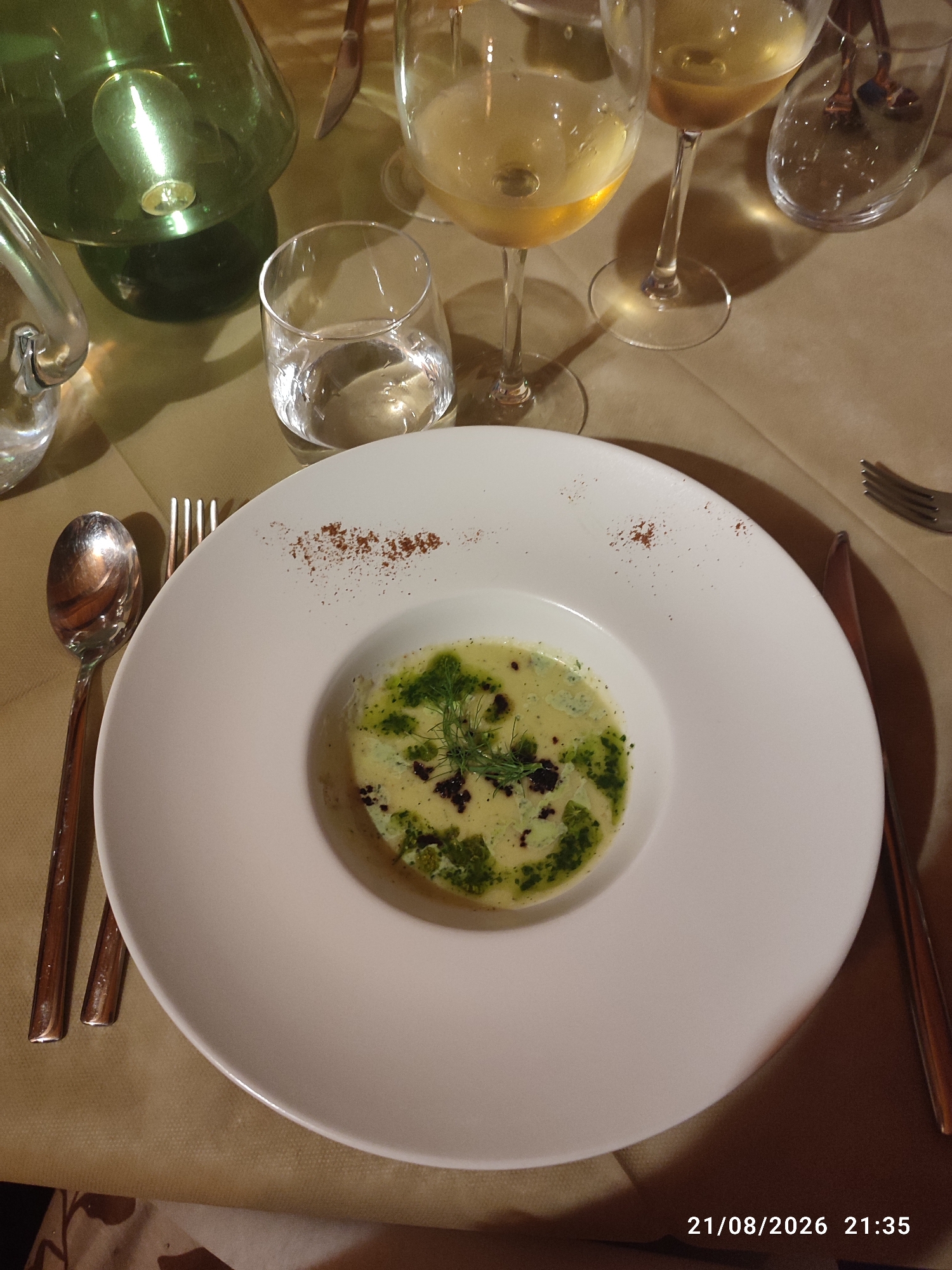 The image shows a close-up of a white plate of food, likely a soup or a creamy dish with green herbs and dark specks, presented in a formal dining setting. The plate is garnished with a light dusting of reddish-brown powder. To the left of the plate, a silver spoon and a fork are visible. In the background, there are two wine glasses filled with a pale yellow liquid, a glass of water, and a dark green carafe. The table is covered with a light brown tablecloth. The lighting suggests an indoor setting, possibly a restaurant, with soft shadows. The timestamp "21/08/2026 21:35" is visible in the bottom right corner.

The location mentioned, Militello in Val di Catania, Italy, is a town in the province of Catania in Sicily. While the image itself does not provide direct visual clues to its specific location within Militello in Val di Catania, it depicts a scene that could be found in a restaurant or home in this Italian town. The overall ambiance suggests a meal being enjoyed, possibly as part of a formal dinner or a special occasion. There are no people visible in the image.