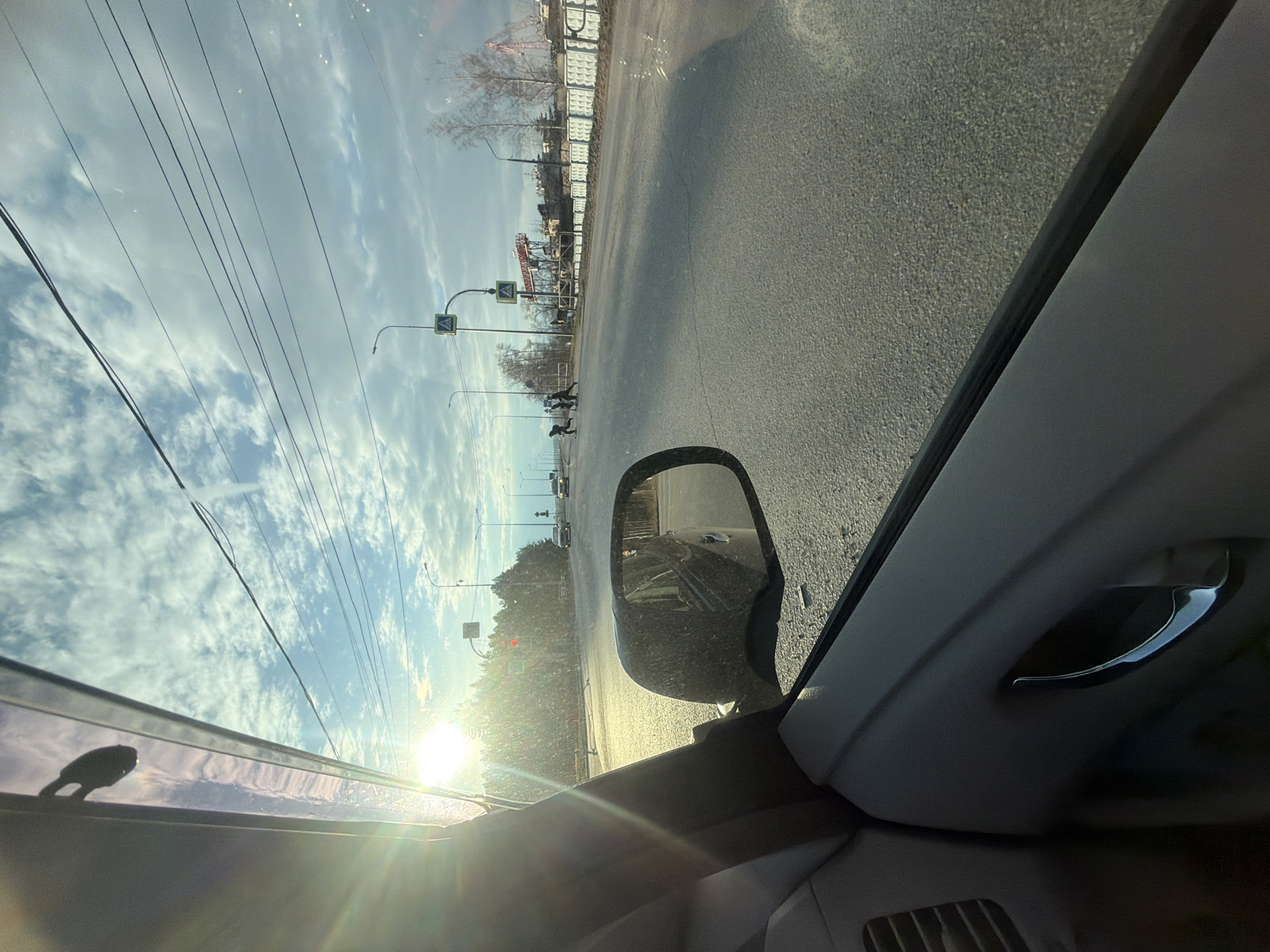 This media file presents a street view from inside a vehicle, likely captured from the passenger seat, looking out at an angle through the side window and windshield. The image is oriented with the car's roofline on the left, suggesting a 90-degree counter-clockwise rotation from a typical upright perspective.
The scene depicts a bright, sunny day with a sky filled with scattered cumulus clouds. A low sun, casting a prominent lens flare in the lower left of the frame (if oriented correctly), suggests either morning or late afternoon. Numerous overhead power lines stretch across the sky.
Outside the car, an asphalt road occupies a significant portion of the view. On the roadside, two yellow diamond-shaped signs with black pedestrian symbols mark a pedestrian crossing, which several individuals are utilizing. Bare trees line the street, indicating a non-summer season. In the background, there is a long, light-colored concrete fence, and industrial or construction structures, including what appears to be a crane. Other vehicles are visible in the distance.
Inside the car, parts of the grey interior are visible, including the door panel, a shiny chrome door handle, and a ventilation grille. The car's side-view mirror reflects the road behind and parts of another vehicle. The strong sunlight creates bright reflections on the car's window and interior surfaces.
The setting is an urban or semi-urban environment in Chaikovsky, Russia. No discernible text is visible, only symbolic traffic signs. The overall impression is of a clear and active day on a city street.