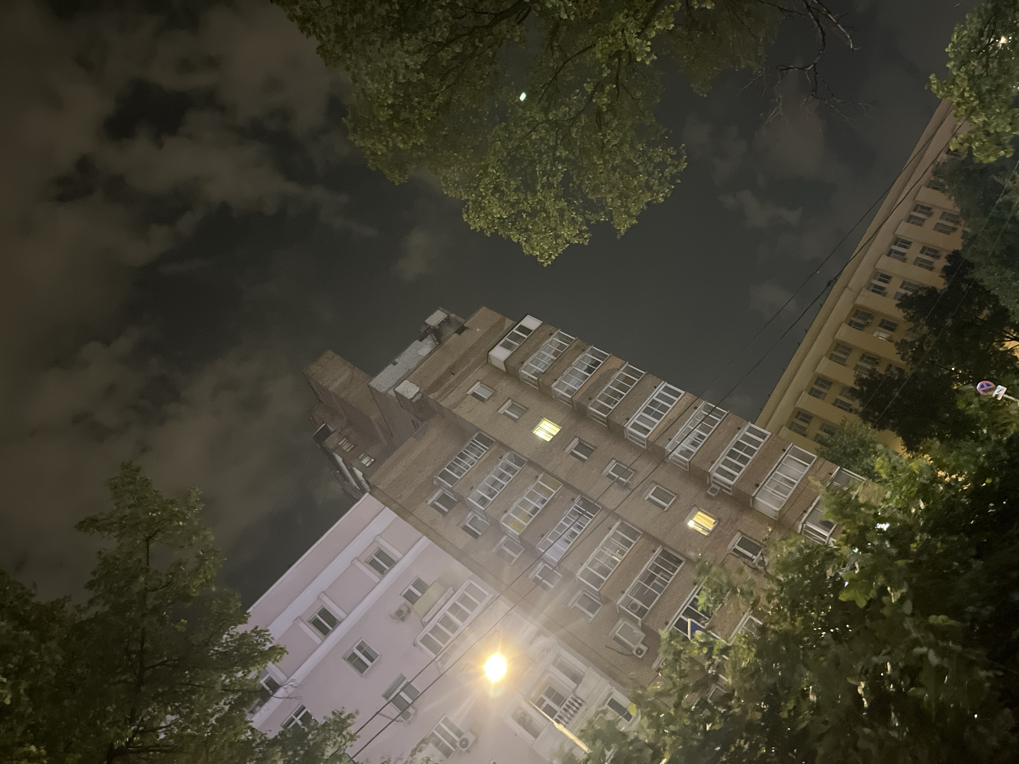 The image shows an upward-angle view of buildings and trees at night, with a cloudy sky. The scene appears to be in a city, with multiple apartment buildings visible. One building is light pink and the other is brick red. A bright street lamp illuminates the area. Trees with green foliage frame the top and sides of the image, partially obscuring the buildings and sky. The overall impression is that of an urban residential area at night, possibly during summer due to the presence of green leaves on the trees. There are no people or specific actions depicted in the image. The context of Moscow, Russia, is not explicitly conveyed by the visual elements alone.