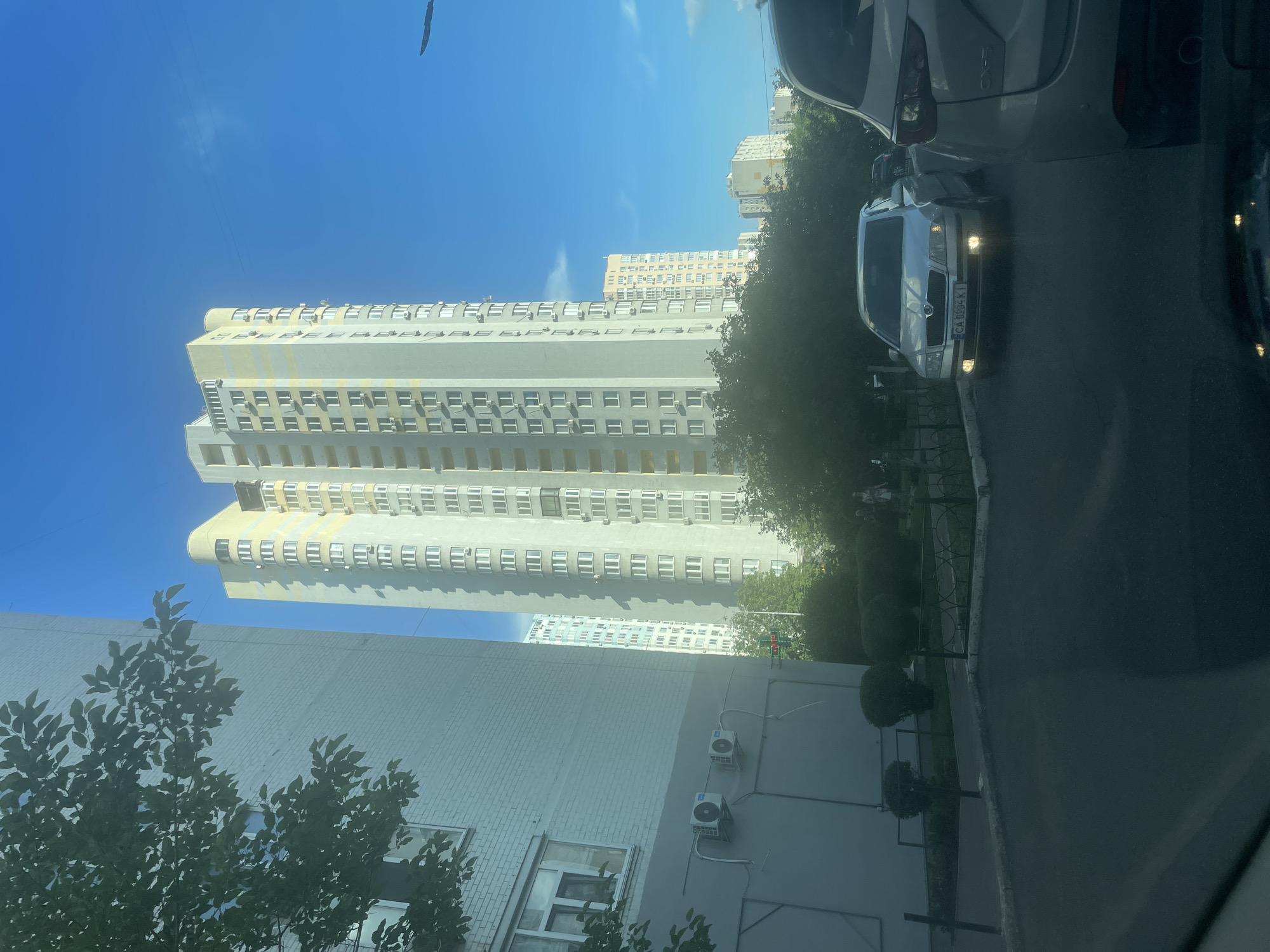 The image depicts a residential street scene in Kyiv, Ukraine, likely during daylight hours under a clear blue sky with sparse clouds. The foreground is dominated by the asphalt road surface and the rear of a white car. To the left, a light-colored multi-story building with a brick facade is visible, featuring windows and two air conditioning units mounted on its exterior wall. Further into the midground, a tall, slender apartment building with a predominantly light gray and pale yellow exterior stands prominently. This building exhibits numerous windows and outdoor air conditioning units along its facade. To the right and behind the central apartment building, other residential structures of varying heights and architectural styles are discernible. Trees and shrubbery line the sidewalk and front yards of the buildings. In the street, a silver Skoda Octavia with a license plate reading "CA 0884 KI" is parked, facing the viewer and with its headlights illuminated. Another vehicle is partially visible behind it. The overall impression is one of a typical urban residential environment.