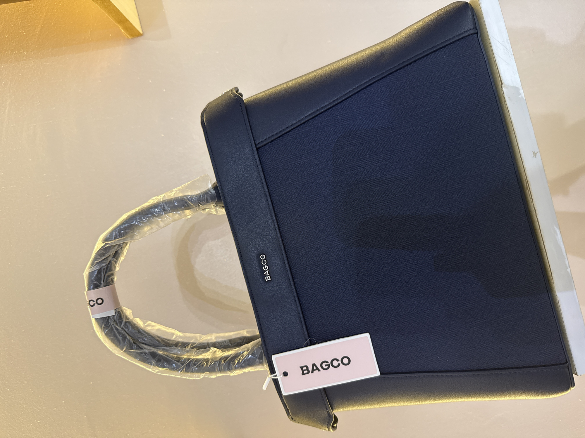 This media file showcases a dark blue BAGCO brand handbag. The handbag is the primary object of focus, presented from a slightly overhead angle. It has a structured, rectangular shape with a textured surface that appears to be a herringbone pattern. A solid dark blue leather trim frames the textured panel, and a dark blue leather handle is visible, partially covered in clear plastic protective wrap.
Attached to the handbag is a pale pink rectangular tag with the text "BAGCO" in dark, sans-serif font. Another small band, also with "CO" visible, is wrapped around the plastic-covered handle. A small silver logo or emblem with the text "BAGCO" is also embossed on the leather trim of the handbag itself.
The setting appears to be indoors, with a light-colored, possibly textured, surface beneath the bag. A sliver of a light-colored, possibly wooden, object is visible in the upper left corner, and a white surface with some scuff marks is present on the right edge of the frame. The lighting is soft and appears to be artificial, suggesting it could be daytime or evening indoors. There are no people visible in the media file. The overall impression is that of a product photograph, possibly for retail or promotional purposes, highlighting the design and branding of the handbag. No specific time of day, weather, or emotional cues are evident. The location context of Abuja, Nigeria, is not directly discernible from visual cues within the image itself but is provided as external information.