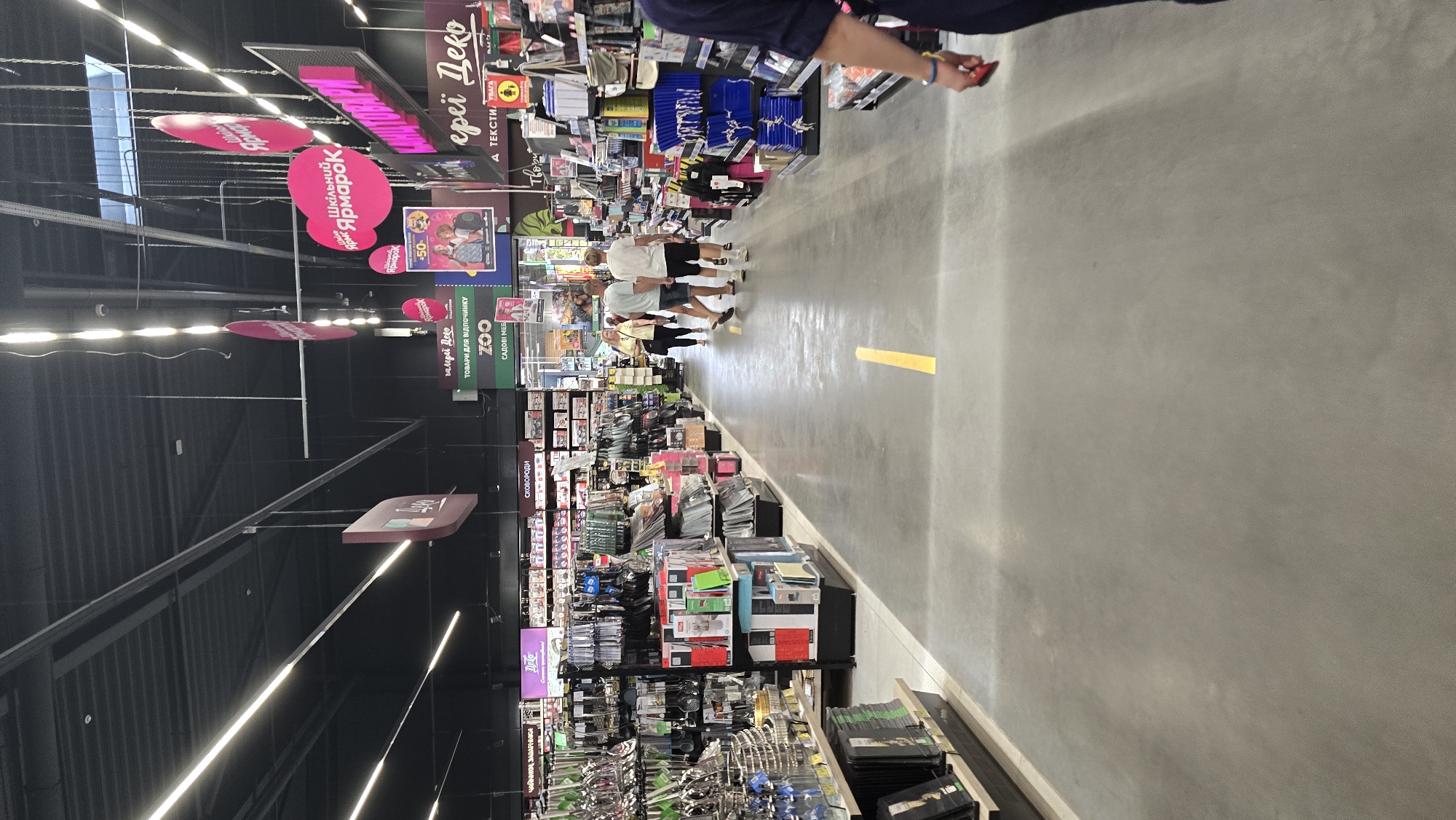 The image depicts the interior of a large retail store, likely a home goods or general merchandise store, with aisles of products visible. In the foreground, a person's arm and torso are visible on the right, wearing a dark blue shirt and holding a red object. The floor is a concrete expanse with a yellow line painted on it, indicating a walkway or lane. In the mid-ground, several shoppers are walking down the aisle. On the left, shelves are stocked with kitchenware, including pots, pans, and utensils. On the right, shelves display various boxed items and packaged goods. Overhead, the store features a dark ceiling with extensive lighting fixtures and hanging signage. One prominent sign in Ukrainian reads "Шкільний Ярмарок" (School Fair), indicating a seasonal sale or product category. Other signs in Ukrainian are visible, such as "Галерея Деко" (Gallery Deco) and "ТОВАРИ ДЛЯ ВІДПОЧИНКУ" (Leisure Goods), suggesting the store offers a wide range of home and lifestyle products. The overall impression is a well-lit, organized retail environment. The presence of Ukrainian signage strongly suggests the location is in Ukraine. Given the specific signage and general context of retail stores, this image is likely from the "Silpo" supermarket chain, a prominent Ukrainian retailer that often includes a "Gallery Deco" section for home goods. While the image itself doesn't definitively pinpoint Zhytomyr, it is highly probable that this scene is from a Silpo store within Zhytomyr, Ukraine, given the commonality of such establishments in Ukrainian cities and the linguistic cues.