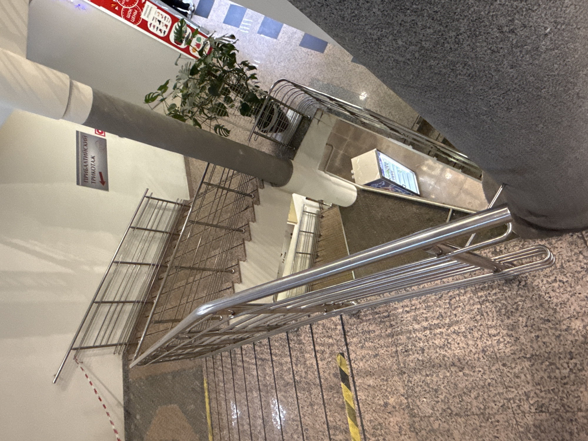 The image depicts an indoor staircase with metal railings. The stairs are covered in brown and grey speckled tile. Yellow and black safety tape marks the edges of some steps. A grey column supports the structure, with a sign reading "ПРИБАЛТИЙСКИЙ ТРИКОТАЖ" (Baltic Knitwear) in Russian. A potted plant sits on a landing near the top of the stairs. The location appears to be a commercial building or shopping center in Moscow, Russia, based on the Cyrillic signage.