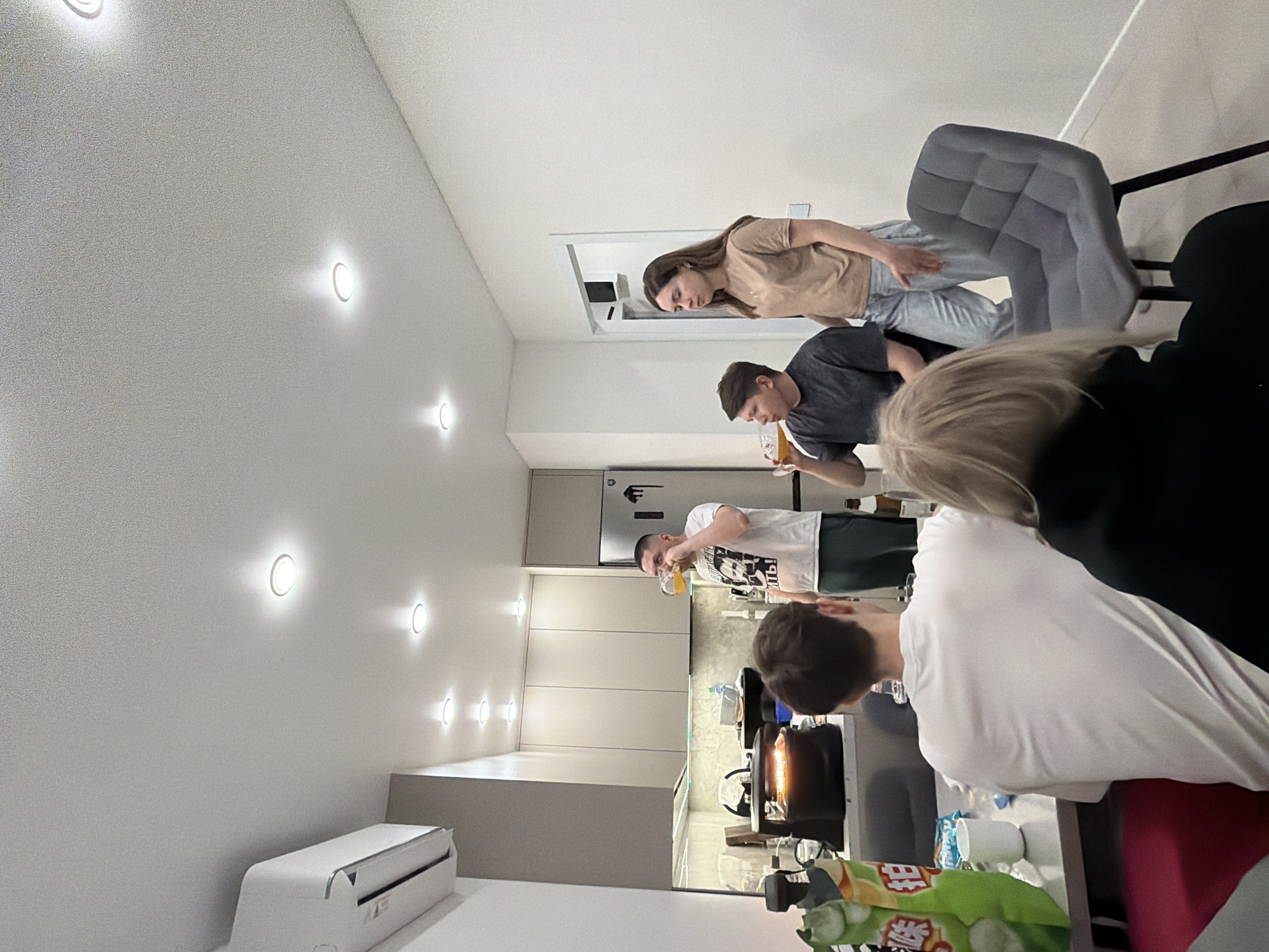 In an indoor setting that appears to be a modern apartment, four individuals are present, seemingly engaged in casual activity. A woman stands to the left of the frame, wearing a beige top and light-wash jeans. Nearby, a man in a dark gray t-shirt is drinking from a glass. In the background, another man, clad in a white t-shirt and dark green shorts, is also drinking from a glass. In the foreground, another individual in a white shirt is also visible.
The setting is a well-lit kitchen, equipped with white cabinetry, a gray-colored wall, and a visible refrigerator. In the lower part of the image, a table surface is visible with various items. A light gray chair is also within the scene. The indoor air conditioning unit is visible mounted to the ceiling.
There is no contextual information within the image to confirm the location as Izhevsk, Russia.