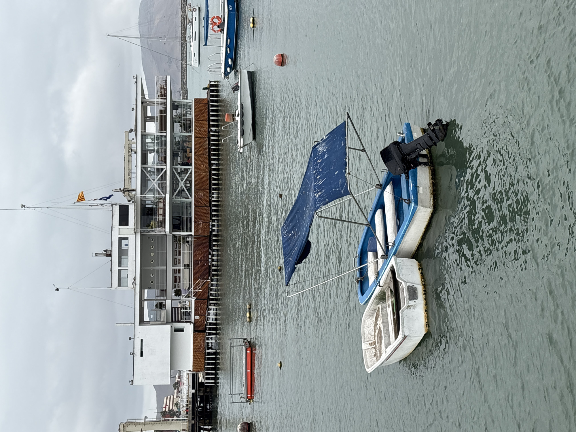 The image depicts a cloudy day at the port of La Punta, Peru. In the foreground, two small boats are moored in the water. The boat on the left is white and appears empty. The boat on the right is blue and white and has a blue canopy. It is equipped with a black outboard motor. In the midground, a large, multi-story white building with extensive glass windows stands on stilts over the water. A pier extends from the left side of the frame to this building. Several other boats and buoys are visible in the water in the midground and background, including a white sailboat with the registration "AYO." The background shows a hilly coastline under a gray, overcast sky.