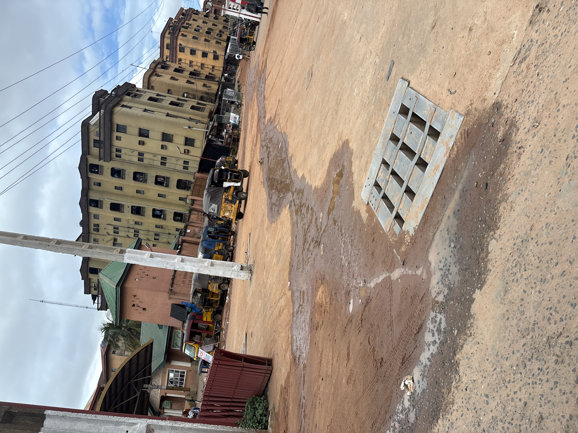 The image shows a street scene in Nkpor, Nigeria. The ground is a mix of red mud and asphalt, with several puddles of water. In the foreground, a metal grate covers a drainage opening. In the mid-ground, several auto-rickshaws, also known as "keke napep," are parked. Some buildings are visible in the background, including multi-story structures with balconies and a building with a green roof. Power lines crisscross the sky. The sky is overcast with some patches of blue. There are a few individuals visible in the scene, some standing near the auto-rickshaws and one walking away from the camera. The overall impression is of a busy, somewhat unpaved urban environment.