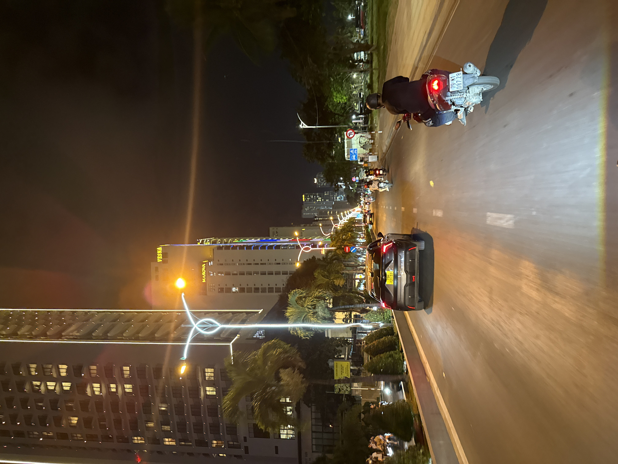 This is a nighttime, vertical, wide-angle shot of a street in Nha Trang Ward, Vietnam. The scene depicts urban nightlife with illuminated buildings, streetlights, and vehicles. In the foreground, a motorcycle with a rider is moving away from the camera on the right side of the road. The motorcycle's license plate is partially visible, reading "79-M1 087.28". Further down the road, a black car is parked or moving slowly on the left. The car's license plate is also visible, reading "19H 121.57".
The background shows a city skyline with several tall buildings. One building on the left has many illuminated windows, suggesting it's an apartment or hotel. Another building on the right has "VESNA" and "MASOVA" written in illuminated letters. Palm trees are scattered throughout the scene, adding to the tropical atmosphere. The streetlights and building lights cast a warm glow on the wet asphalt, indicating recent rain or high humidity. The overall mood is vibrant and active, characteristic of a bustling city at night. The time of day is clearly night due to the darkness and artificial lighting.