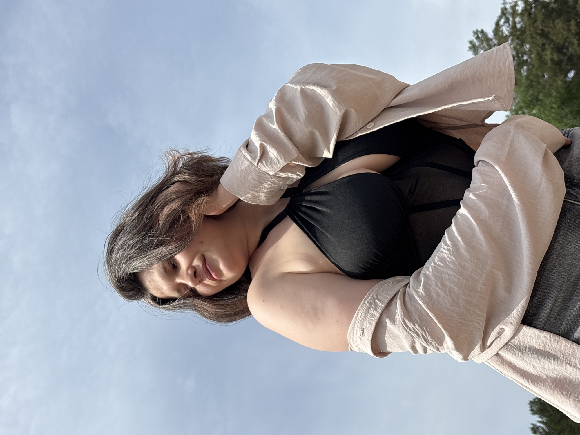 A woman with brown hair and gray streaks poses outdoors, looking downward and to her left. She wears a black top and a light beige shirt. The sky is light blue, and some green foliage is visible in the upper right corner. The location is unspecified, but the prompt suggests it is in the city of Chaikovsky, Russia.