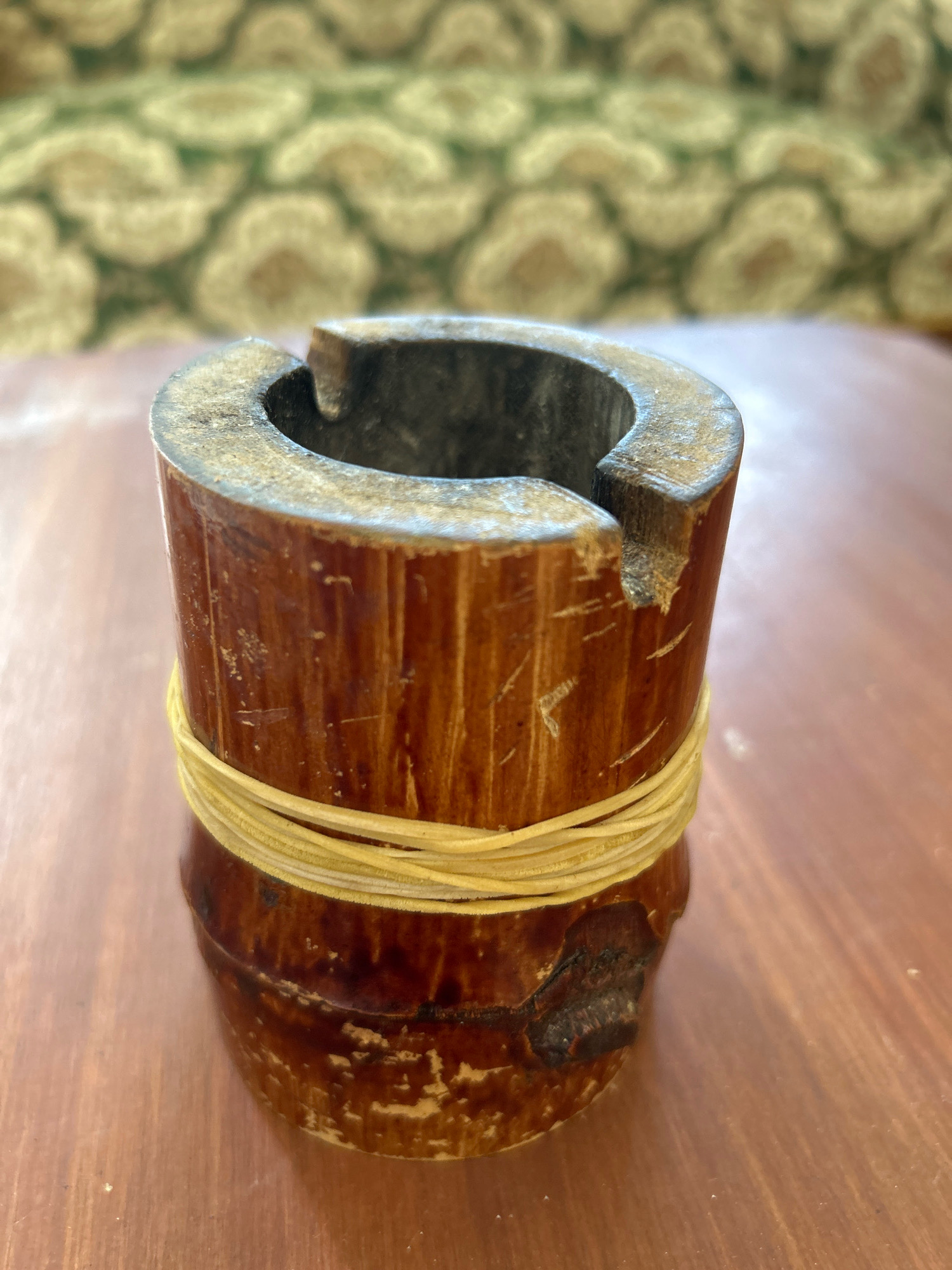 A close-up, eye-level shot captures a rustic, cylindrical wooden ash receptacle resting on a polished wooden table. The ash receptacle, approximately 4 inches tall and 3 inches in diameter, is crafted from dark brown wood with visible grain patterns and signs of wear, including nicks and scratches. A bundle of thin, light yellow elastic bands encircles the middle of the receptacle. The top of the receptacle is hollowed out, forming a small bowl with a notched opening, typical of an ashtray. The background is softly blurred, revealing a patterned upholstery in shades of green and cream, likely a couch or chair. The lighting suggests an indoor setting, possibly a cafe or a home in the city of Sabang, Indonesia, given the implied context.
