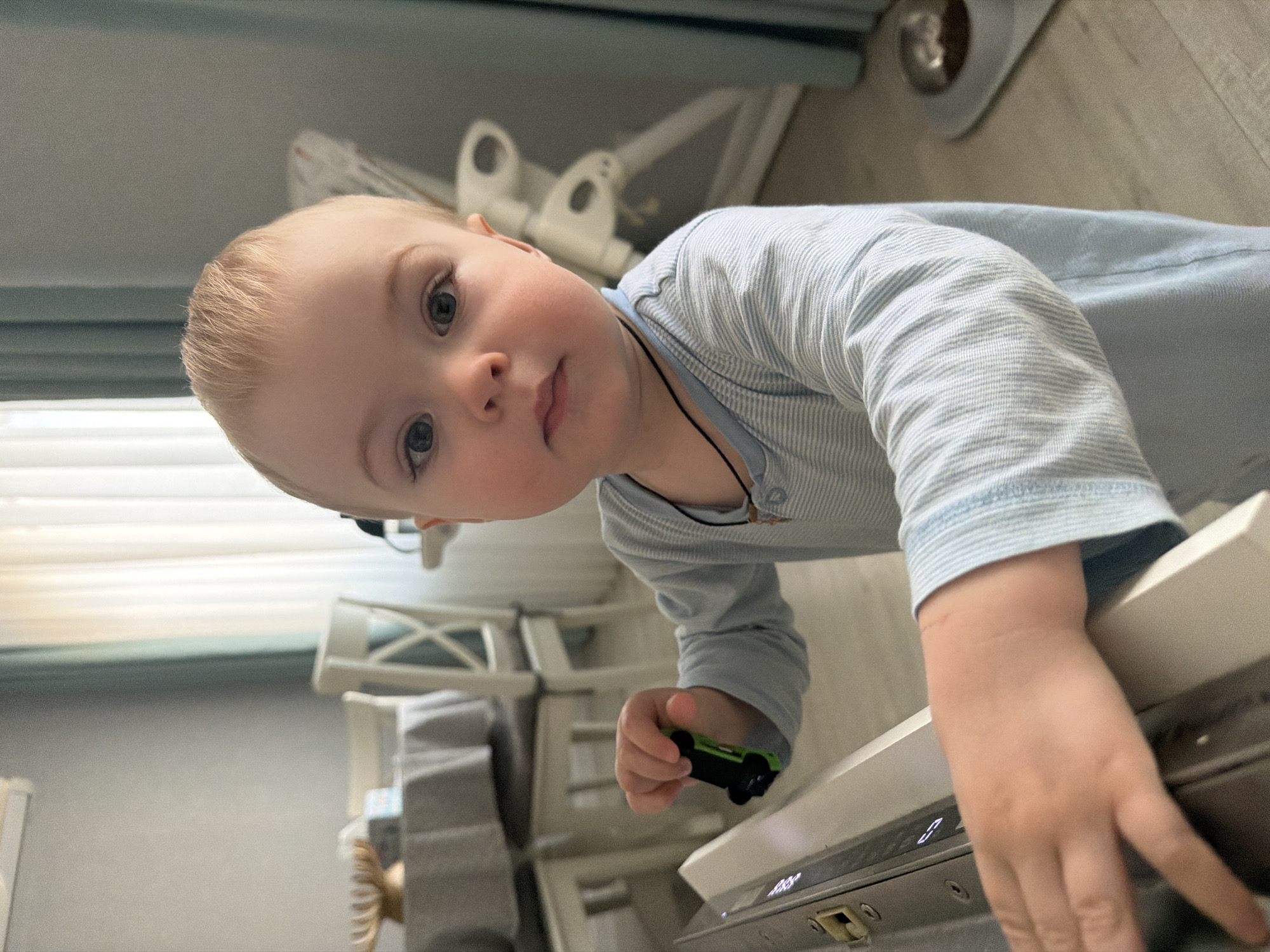 A young child with blond hair and light eyes is leaning over a kitchen counter, looking at the camera. The child is wearing a light blue striped long-sleeved shirt and has a thin necklace with a pendant. The child holds a small green and black toy car in one hand. The counter appears to be part of a kitchen appliance. The background shows a partially visible window with a curtain, a child's high chair, and a glimpse of a floor with what looks like a metal dog bowl. The location is presumably a home setting in Votkinsk, Russia, but there are no details in the image to definitively confirm it.