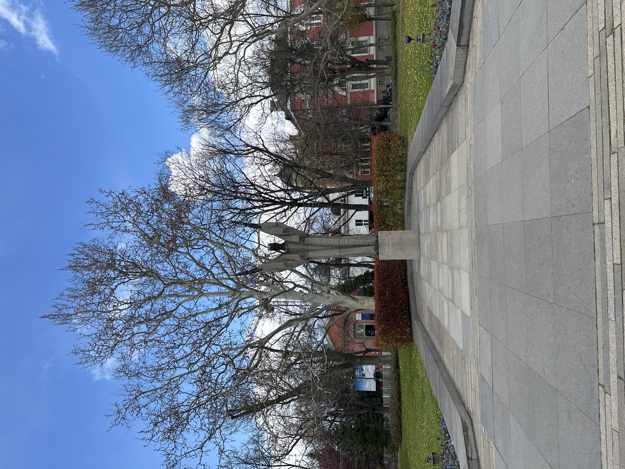 The image shows a park scene in Sofia, Bulgaria, likely near the National Palace of Culture (NDK). A stone monument or memorial stands in the foreground, partially obscured by a large bare tree. Behind the monument is a row of bushes, a green lawn dotted with yellow flowers, and a red brick building. In the distance, people are gathered near the building. The sky is blue with scattered white clouds. A paved walkway extends from the lower right corner into the scene.