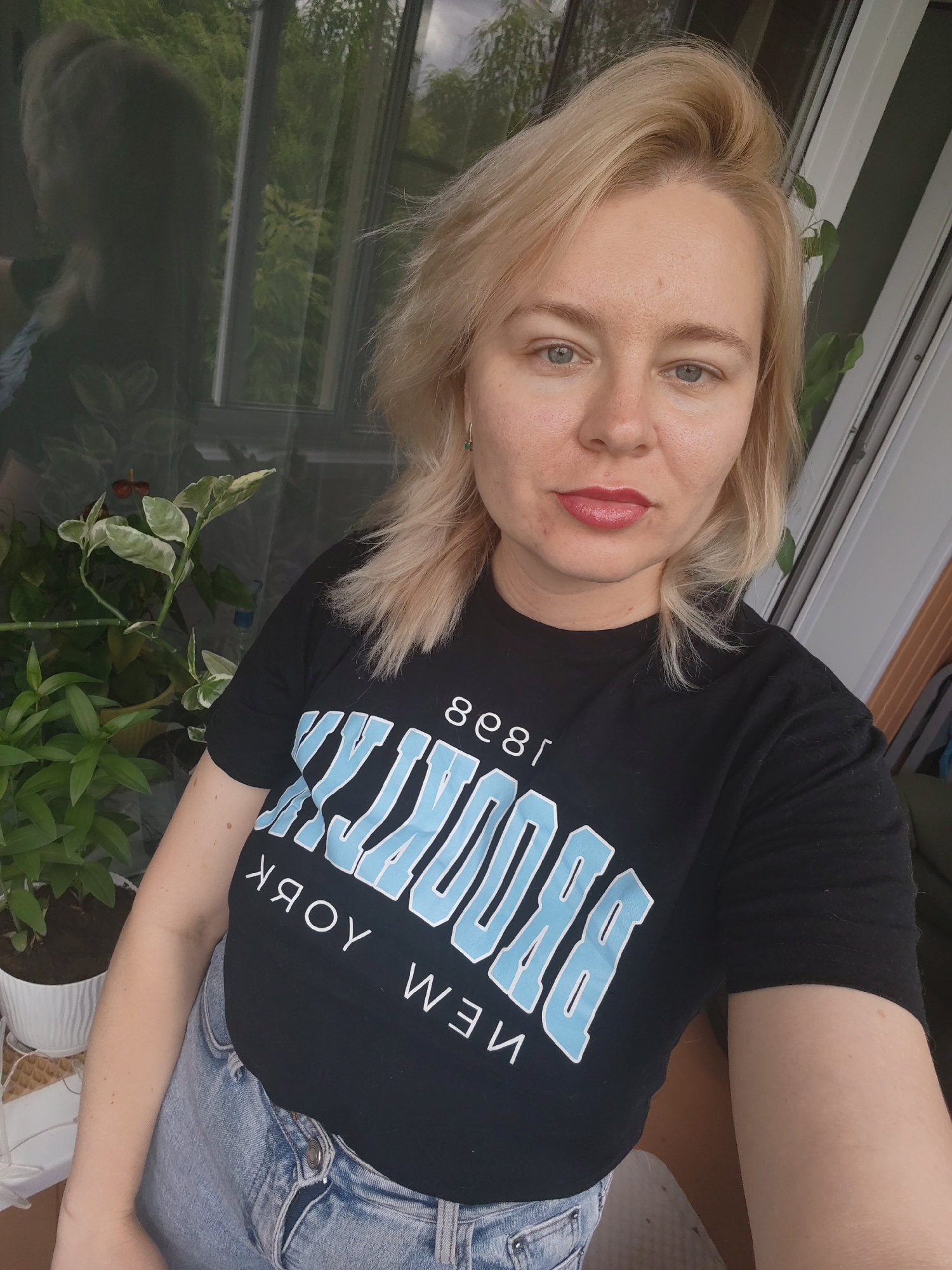 A Caucasian woman with blonde hair and light blue eyes is pictured in a selfie. She is wearing a black t-shirt with light blue lettering that says "BROOKLYN" and "NEW YORK" and light blue denim jeans. She has on red lipstick and appears to be indoors, possibly on a balcony, with plants visible behind her and a reflection of trees in a window or glass door. The image was captured on June 27, 2023, at 2:36 PM. The location of Izhevsk, Russia, is provided as context, but there are no specific visual cues within the image to confirm this location.