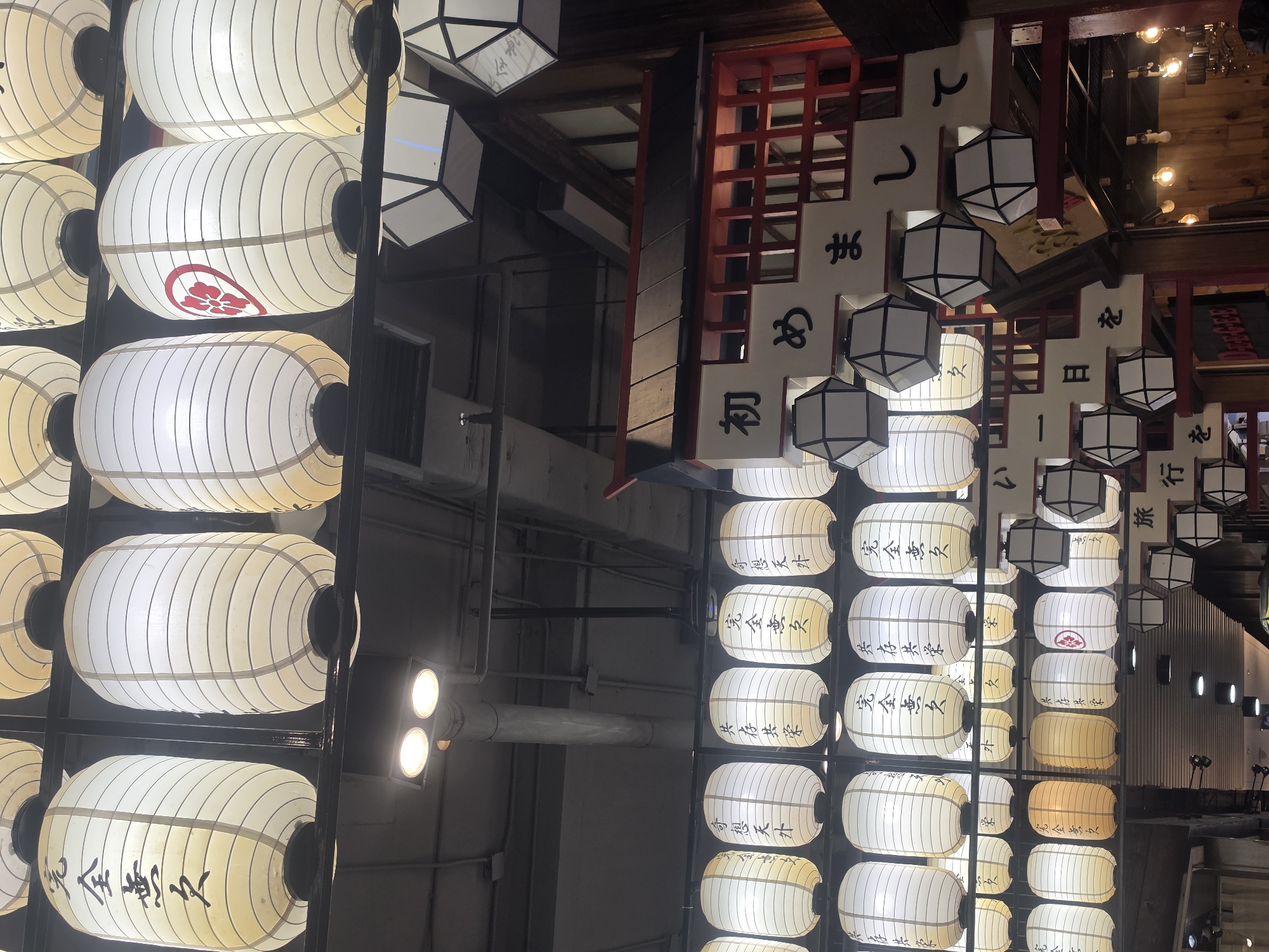 The image captures an indoor setting in Bangkok, Thailand, as indicated by the presence of traditional Japanese lanterns and signage in Japanese characters. Two prominent arrays of lanterns dominate the left and right sides of the frame. The lanterns on the left, uniformly white, are arranged vertically in a grid, some with printed characters or symbols. In contrast, the lanterns on the right display a gradient of colors, transitioning from white at the top to a warm yellow towards the bottom.
Additional elements in the scene include red structural support beams, wooden elements, and hanging white signs with Japanese characters, interspersed with cube-shaped paper lanterns. Background lighting fixtures and pipes are visible, contributing to the environment. The composition suggests a space combining aspects of Japanese cultural aesthetics within the unique setting of Bangkok.