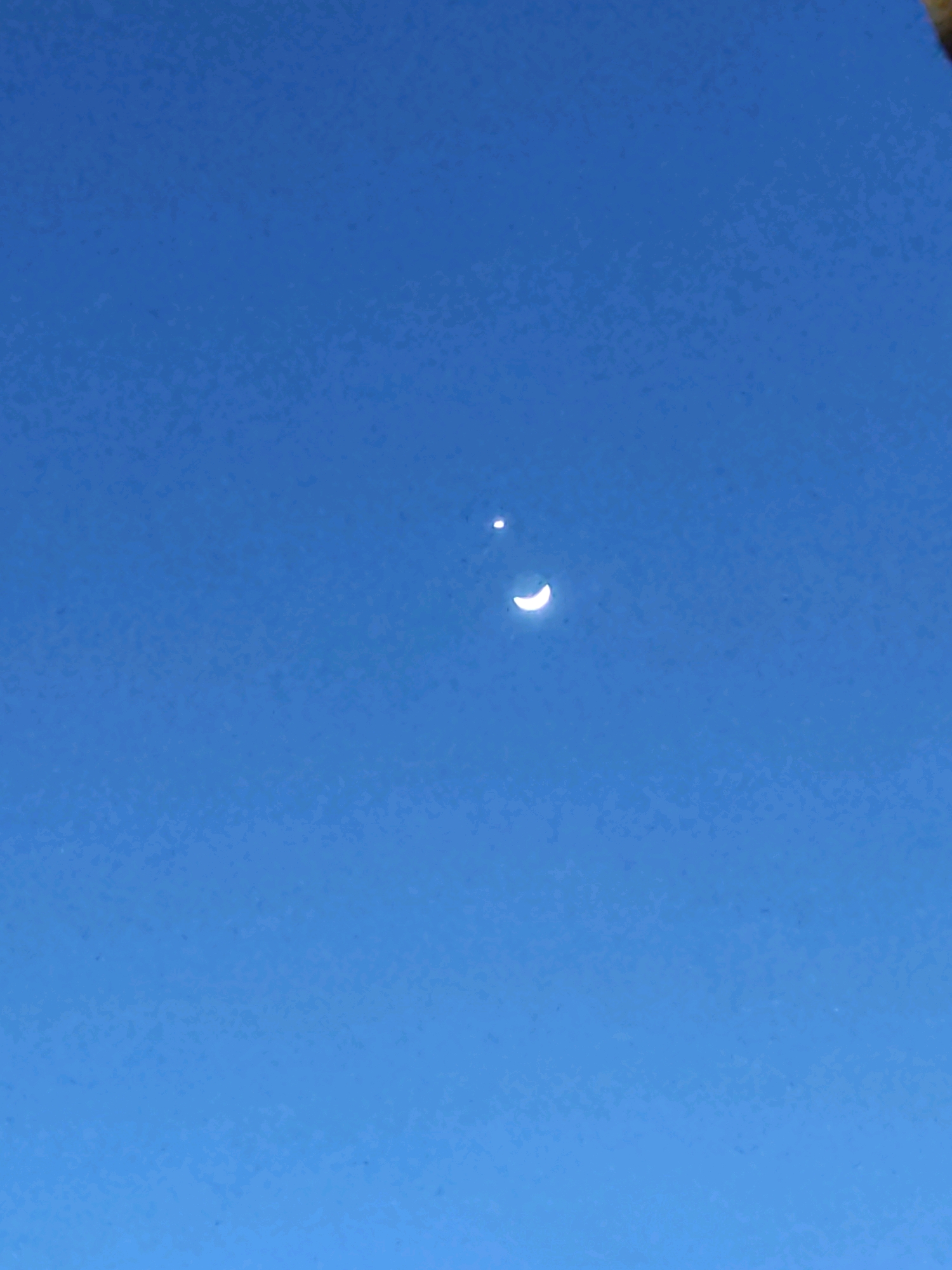 The image displays a clear blue sky with two celestial bodies. A crescent moon, illuminated on its right side, is visible in the lower center of the frame. Above and slightly to the left of the moon, a smaller, bright circular object is present. The sky exhibits a uniform blue hue, suggesting daytime or twilight conditions. The location is identified as Tiudan, Indonesia.