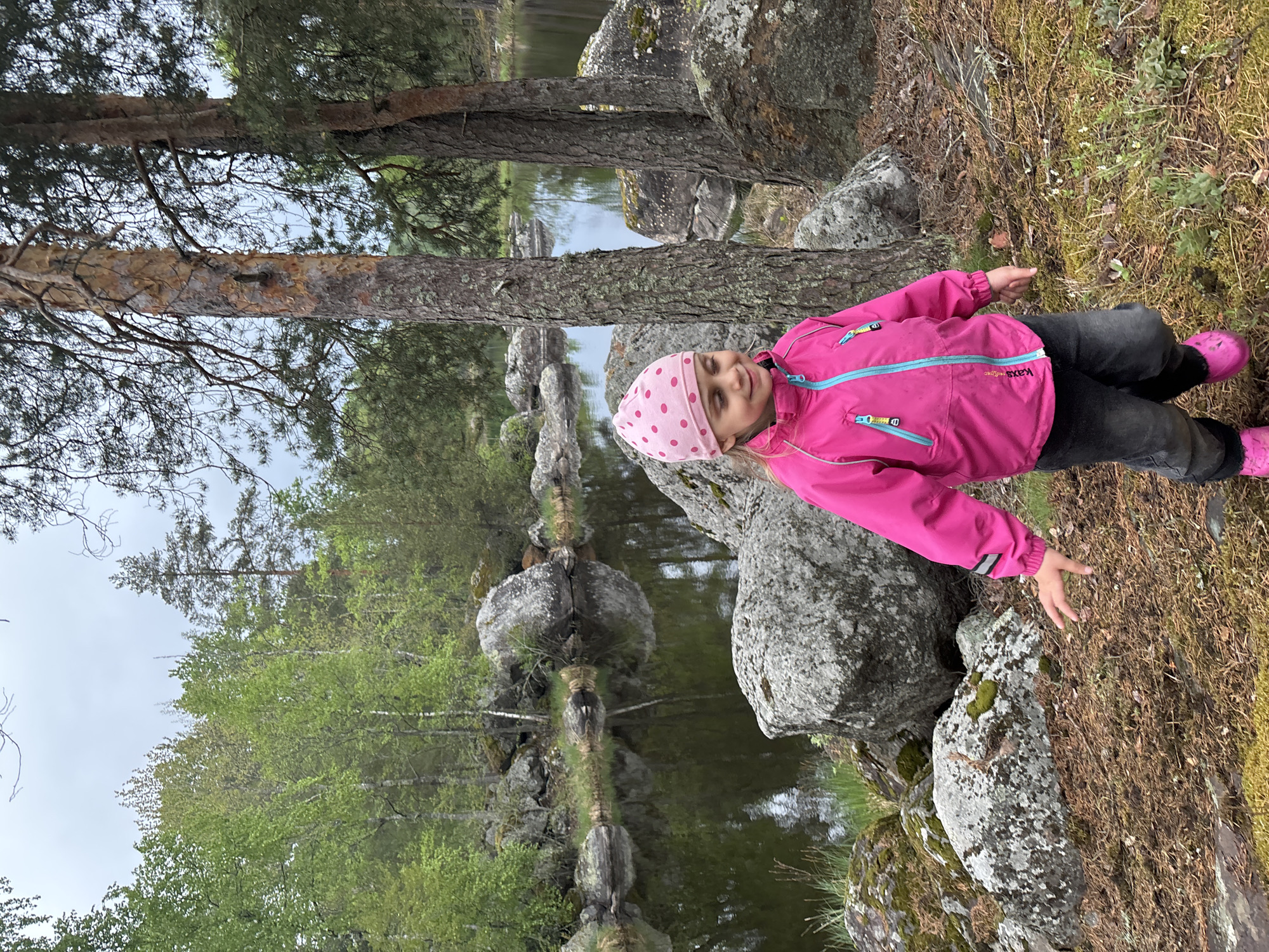 The image shows a young girl outdoors, possibly near a body of water. She is wearing a bright pink jacket, dark pants, a pink hat with polka dots, and pink shoes. The girl stands among large gray rocks covered with moss and some grassy, wooded ground. Behind her is a body of water with reflections of trees and rocks. The trees in the background are a mix of deciduous trees with light green leaves and coniferous trees. The sky appears overcast. The location is unknown.