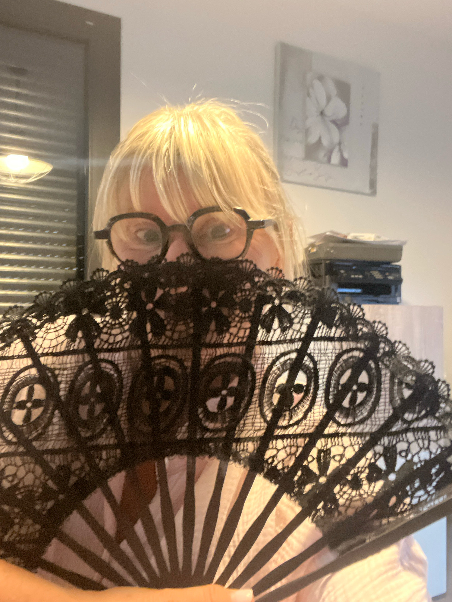 A woman with blonde hair and glasses is holding a black lace fan in front of her face, obscuring her mouth and lower face. She is wearing a light-colored shirt. The background appears to be an indoor setting with blinds on the left and a framed picture of a flower on the right. A piece of office equipment, possibly a printer or scanner, is visible on a shelf behind her. The image was captured in Chambœuf, France.