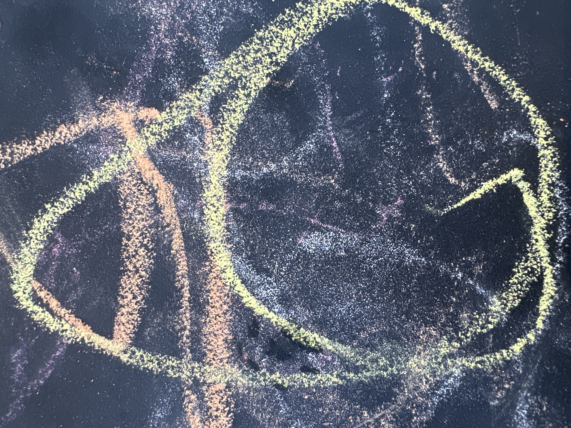 The image is a close-up, high-angle shot of a dark surface, likely a chalkboard or dark paper, with colorful chalk markings. The chalk lines are primarily in shades of yellow, pinkish-orange, and some faint purple and white. The markings appear to be abstract scribbles and shapes, with some thicker, more defined lines forming loops and arcs, and thinner, fainter lines creating a scattered effect. The chalk has a textured, powdery appearance, and there are areas where the chalk dust is more concentrated, giving the impression of a slightly smudged or layered effect. The background is a uniform dark color, showing the texture of the surface. There are no people or recognizable objects in the image. The image does not contain any information that can be used to determine its location within the city of Novoterksky, Russia.
