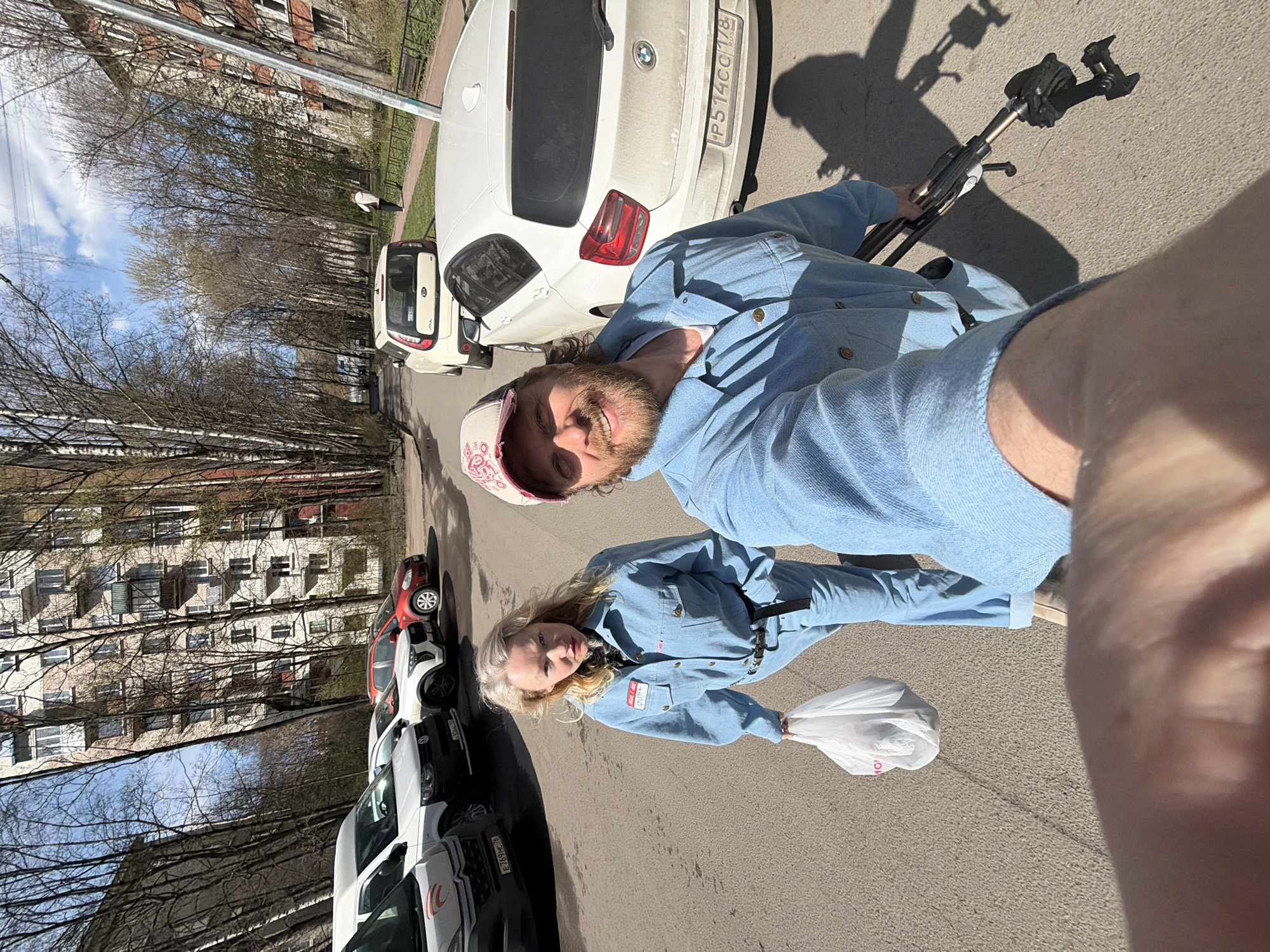 This outdoor selfie captures a man and a woman on a street in Saint Petersburg, Russia, likely during late winter or early spring due to the bare trees. The image is taken under bright, sunny conditions, casting long shadows, suggesting midday or early afternoon.
The man, with a beard and wearing a light blue denim jacket and a baseball cap, smiles broadly towards the camera, which he appears to be holding with a tripod or selfie stick. The woman, with blonde hair and wearing a light blue denim jumpsuit with a black belt, stands beside him with a neutral expression, carrying a white plastic bag. A red patch with the text "PLUS" is visible on her jumpsuit.
They are standing on a paved road, with several parked cars lining the street. Immediately behind them, a white BMW SUV (license plate P514CC 178 RUS) and a white Kia Soul are visible. Further down the street, a red car and other vehicles are parked. In the background, multi-story residential buildings with numerous windows and balconies line the street, partially obscured by the leafless trees. Partially visible Cyrillic text, possibly "ОКО", is on the plastic bag held by the woman.