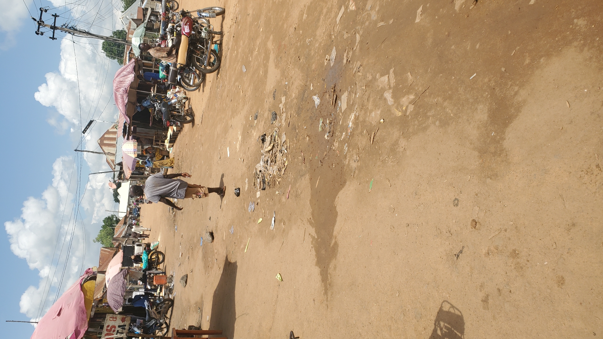 The image depicts a bustling outdoor market scene in Jalingo, Nigeria. A person, wearing a grey t-shirt and brown shorts, walks away from the camera in the foreground. The ground is predominantly dirt with scattered debris, including plastic bags, paper, and what appears to be a broken ceramic shard. To the left of the frame, market stalls are set up with colorful tarps, one pink and one yellow, offering goods. Several motorcycles are parked or present in the market area. In the mid-ground, more market stalls with various awnings are visible, with people browsing and tending to their wares. A man is seated on a motorcycle in the right mid-ground. The background shows more market structures, utility poles with extensive wiring, and a clear sky with scattered clouds. The overall impression is one of a vibrant, albeit somewhat untidy, local marketplace.