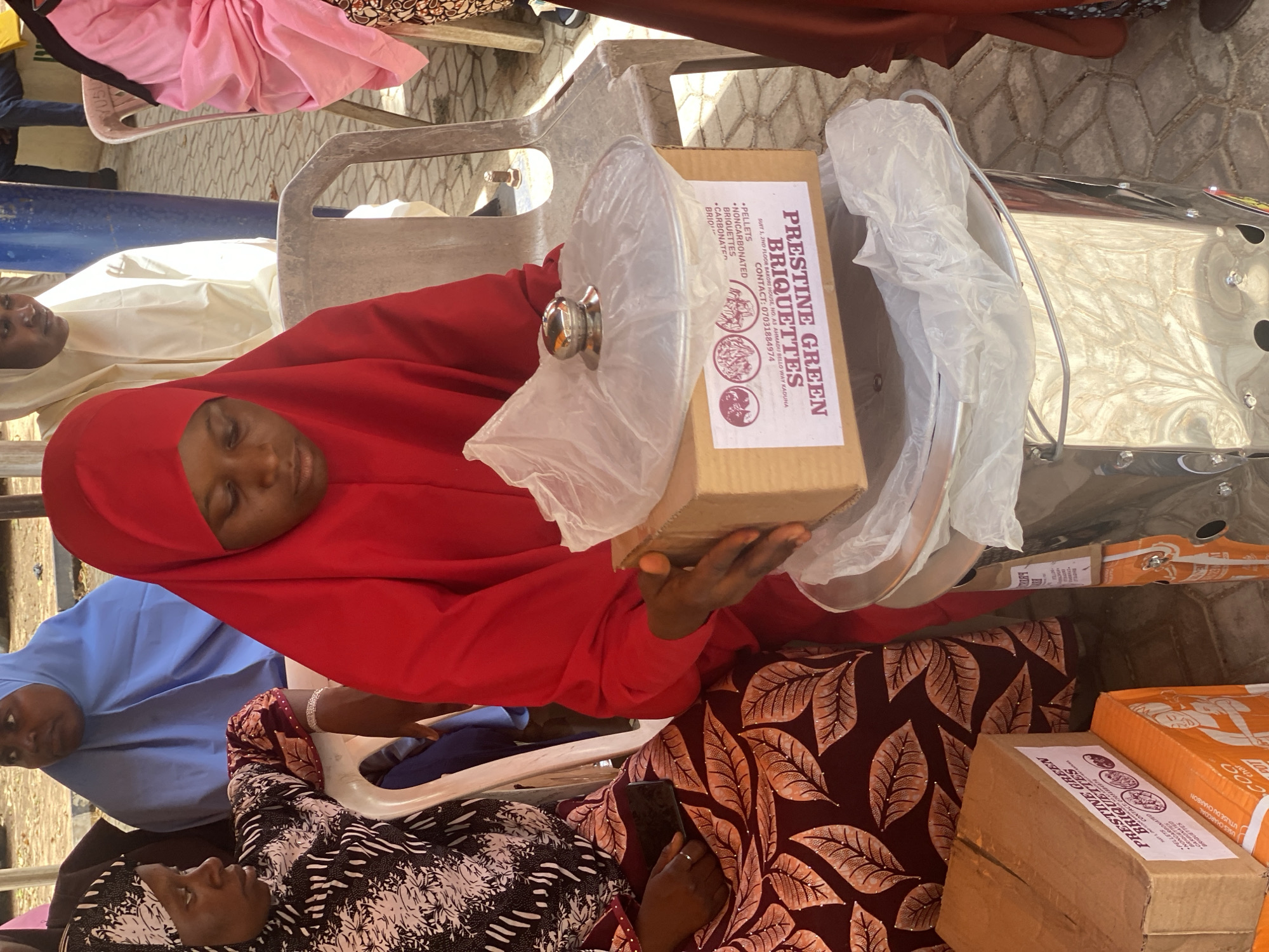 A woman wearing a red hijab is holding a cardboard box labeled "Pristine Green Briquettes" in what appears to be an outdoor setting. The box also lists a Kaduna address: Suit 1, 2nd Floor Bakors House, No. A5 Ahmadu Bello Way Kaduna. The presence of other individuals and boxes suggests this could be a distribution or presentation event. The setting is Unguwar Rimi, Nigeria.