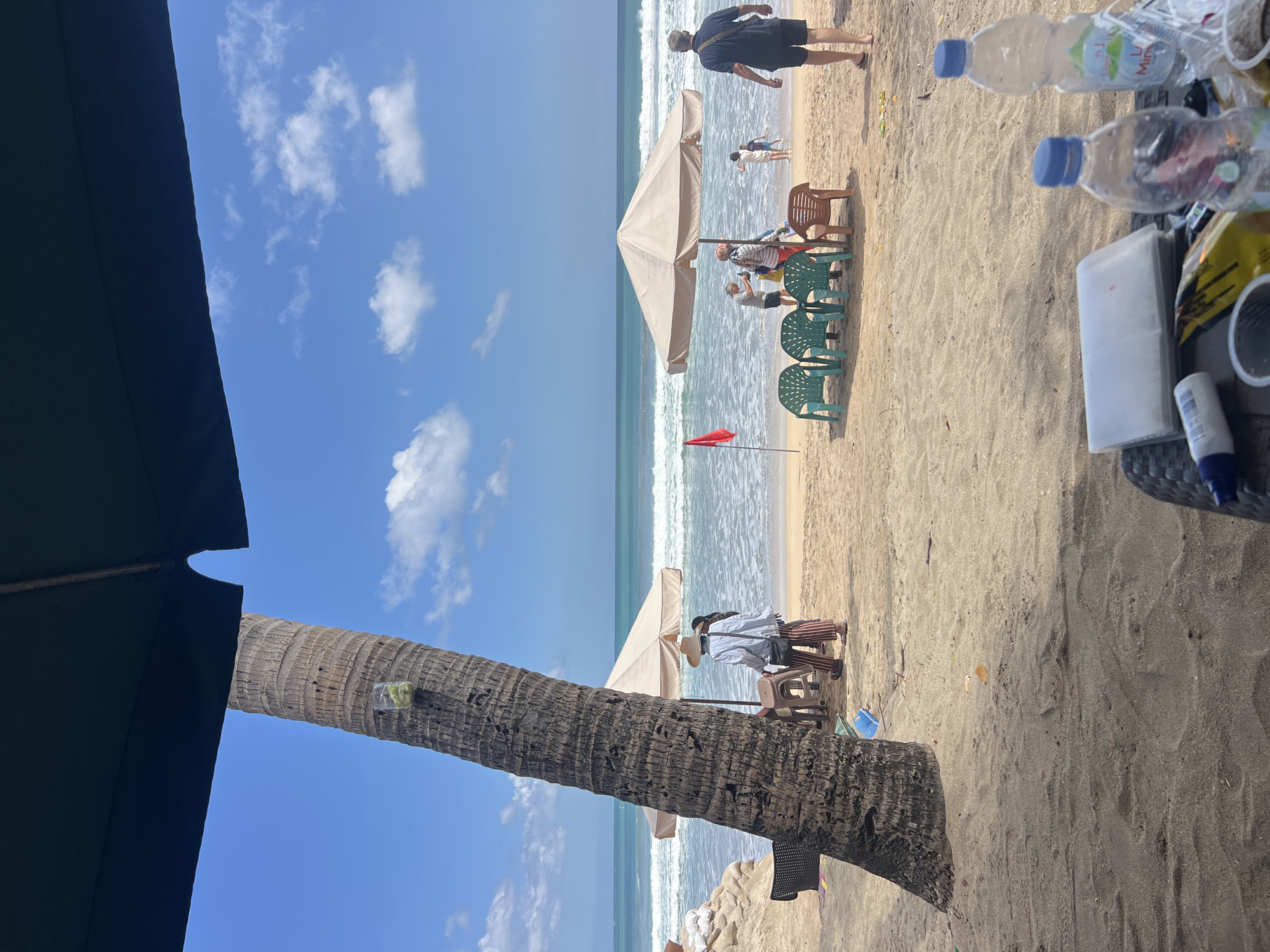 The image depicts a sunny day at Kuta Beach in Bali, Indonesia, with a view of the ocean from a shaded area. In the foreground, a table is laden with water bottles, a small white item, and some snacks. A large palm tree trunk dominates the left side of the frame. Beyond the palm tree, the sandy beach is dotted with beach chairs and umbrellas. Several people are visible on the beach, some walking, others sitting or standing near the water. The ocean is calm with gentle waves, and the sky is blue with scattered white clouds. The overall scene conveys a relaxed atmosphere at a popular tourist destination.