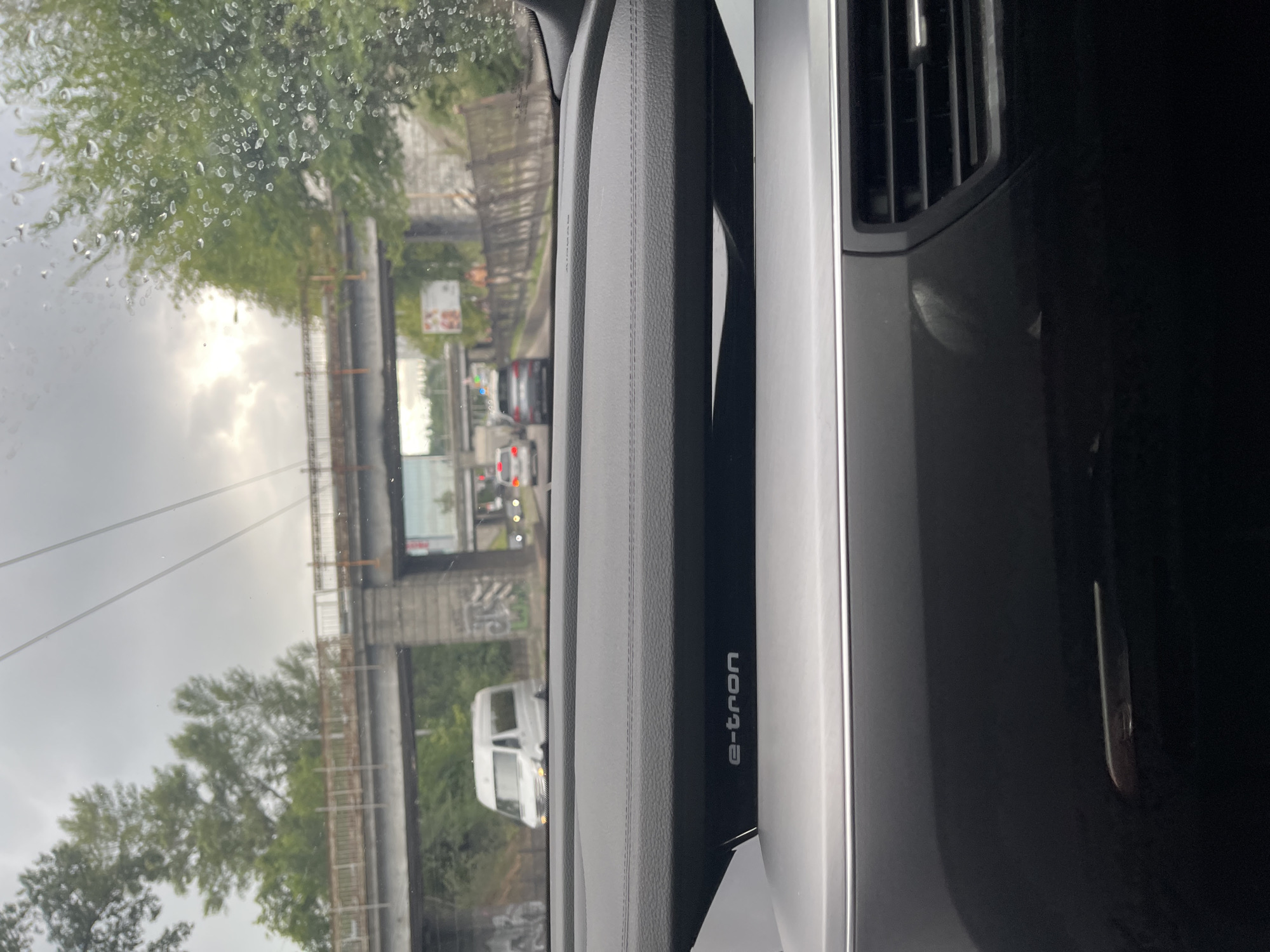 The image is a forward-facing view from inside a vehicle, likely an Audi based on the "e-tron" branding visible on the dashboard. It depicts a road scene under an overcast sky, possibly in Kyiv, Ukraine, given the presence of an overpass structure with graffiti and mature trees lining the roadway. The windshield has raindrops, indicating inclement weather. Traffic is visible on the road, including a white van, a silver sedan, and other vehicles further ahead. An elevated railway or bridge crosses the road in the background. The foreground shows a portion of the car's interior, including the dashboard and air vents.