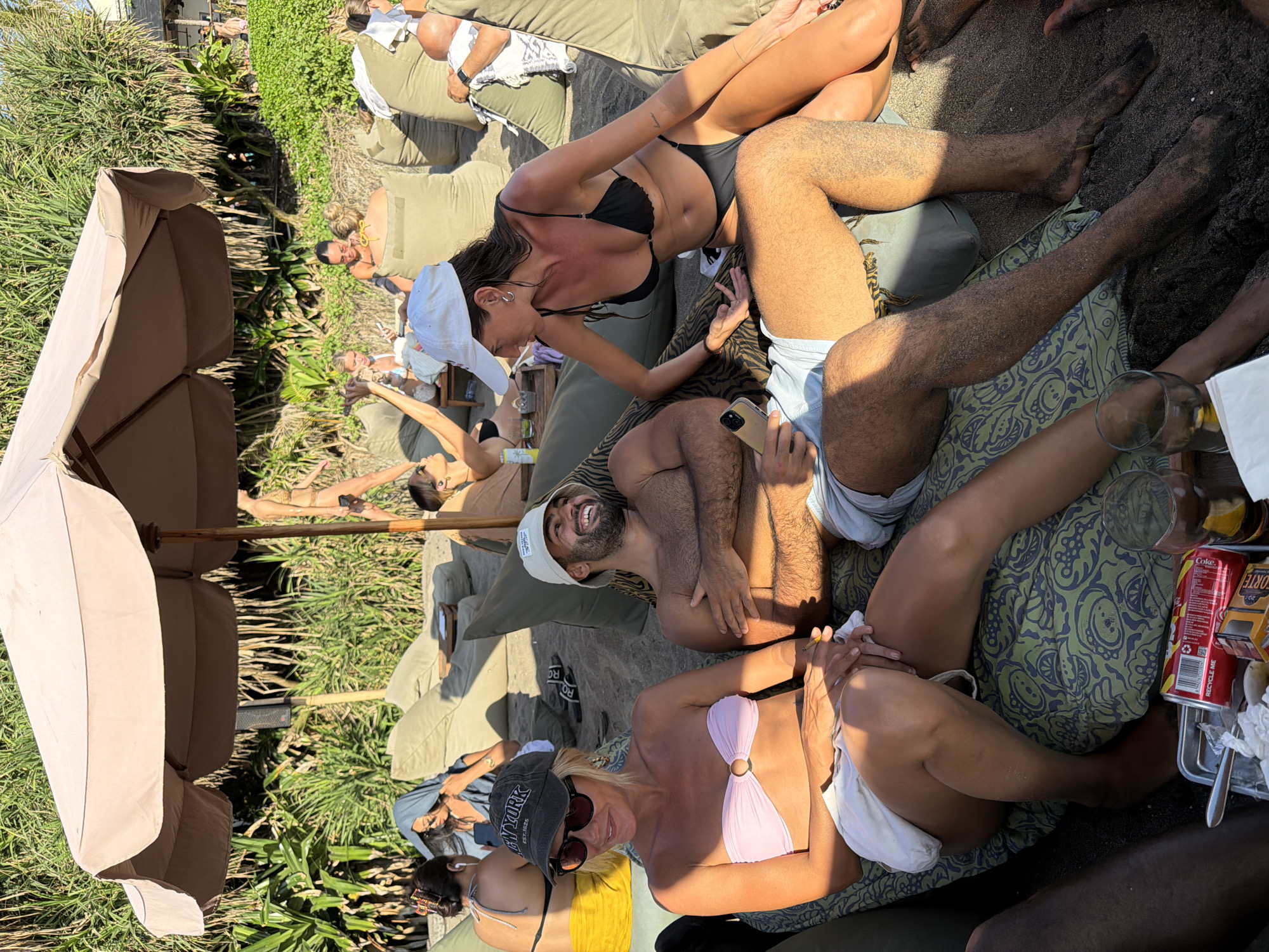 This image shows a group of people relaxing outdoors. Several individuals are lounging on cushions, some wearing swimwear. One person is holding a mobile phone, and others appear to be socializing. A large umbrella provides shade. Drinks and other items are visible on a tray. Based on the presence of black volcanic sand, this scene is likely from one of the beaches in North Kuta, Indonesia.