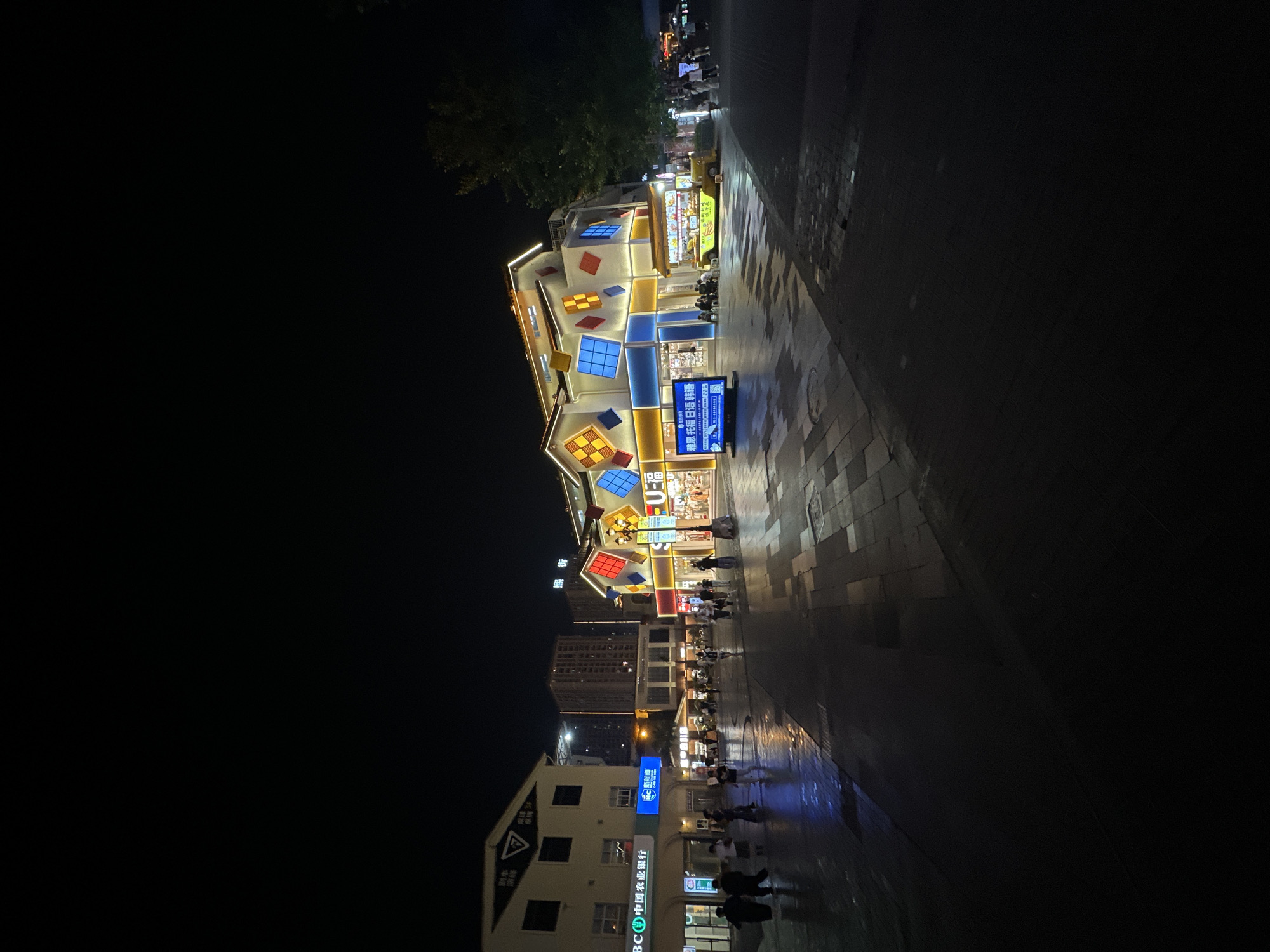 The image depicts a night scene on a street in the Chongqing High-tech Zone, China. A brightly lit building with a colorful diamond-patterned facade is the central focus. Several individuals are walking on the wet, reflective pavement in front of the building and in the distance. To the left, a building with a sign that includes Chinese characters and "BC 中国农业银行" (BC China Agricultural Bank) is visible. A yellow food cart is parked near the brightly lit building. The street is paved with dark tiles, and the reflections on the wet surface indicate recent rain. Tall buildings are visible in the background. The overall atmosphere is that of a lively commercial area at night.