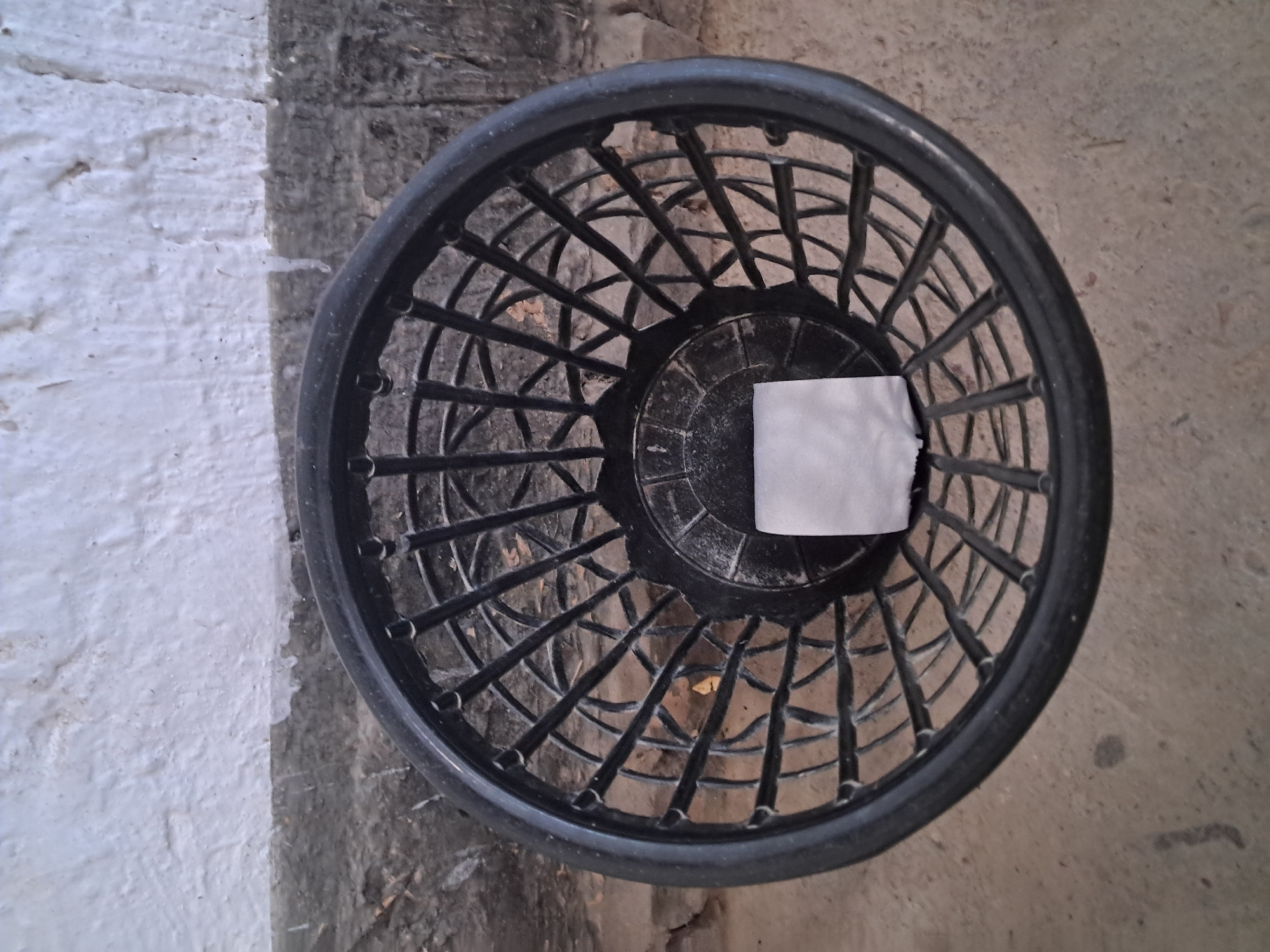 A top-down shot shows a black, woven plastic wastebasket on a concrete floor. The basket contains a single, folded square of white paper. Behind the basket, a rough, textured white wall meets a darker, brick-like surface. The scene appears to be an outdoor or semi-outdoor location in Bagish, Uzbekistan, possibly a public area or a less maintained space.