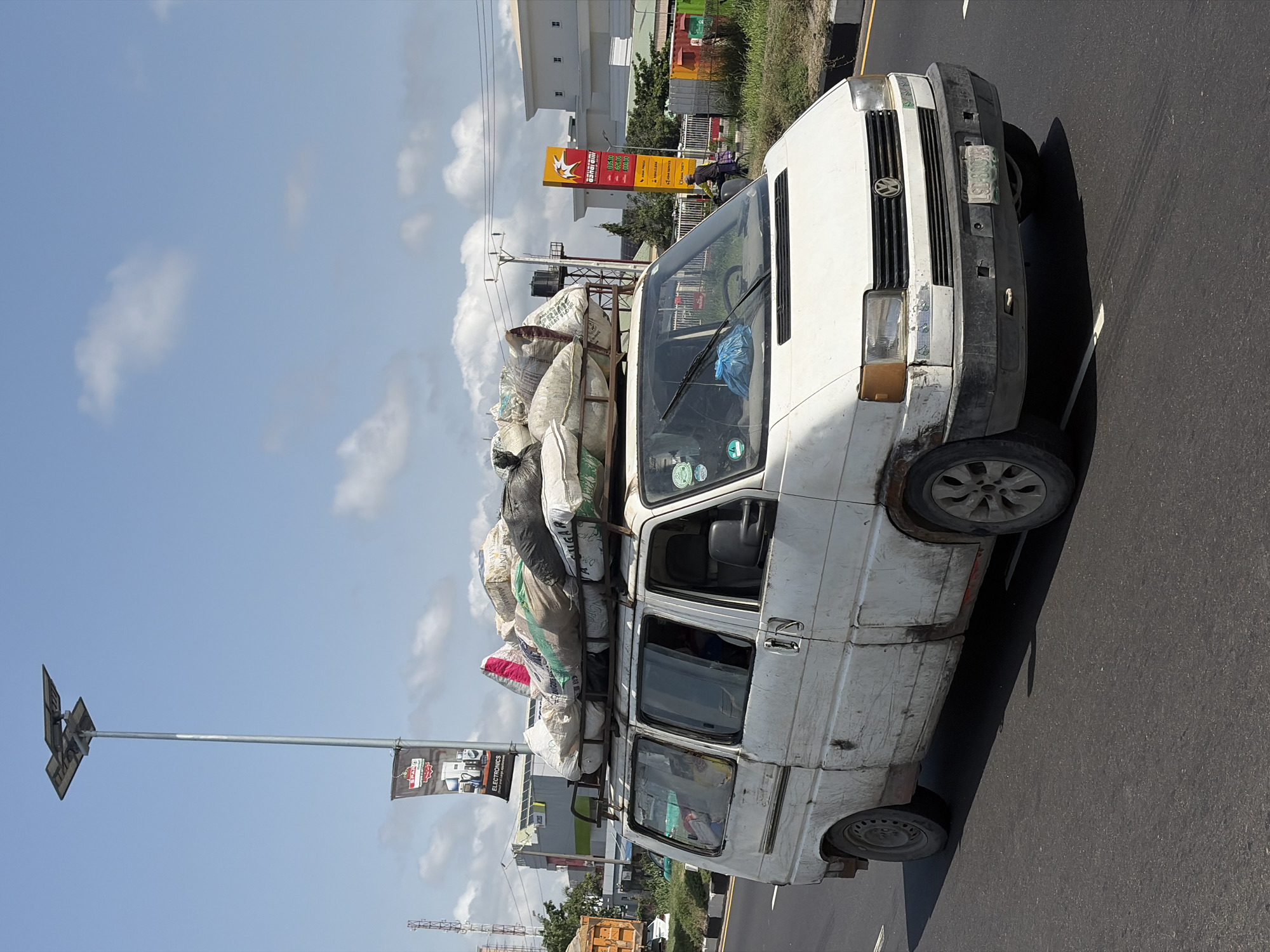 A heavily loaded, white Volkswagen van is driving on a paved road under a clear, bright blue sky with scattered white clouds. The van, appearing old and worn with dirt and some rust, is packed with numerous large white and multi-colored sacks piled high on its roof rack, suggesting it is transporting goods. The front grille displays the Volkswagen logo, and a partially visible Nigerian license plate (S-800K) is seen on the front bumper. Stickers, including one possibly for "LAGOS STATE," are on the windshield.
In the background, on the right side of the road, a yellow and red "ORANGE FUEL" gas station sign is prominent, displaying fuel prices such as "PMS 800.00," "AGO 1000.00," "DPK 850.00," and "LPG 680.00." A person on a bicycle is visible near the gas station. Further back, various buildings, including a large white structure, are seen. Utility poles and power lines crisscross the sky.
On the left side of the road, a tall street light pole with solar panels is visible, from which a banner advertising "SPA... ELECTRONICS" hangs. Green foliage and grass line the roadside. Some of the sacks on the van's roof have visible text, such as "SUGAR" and "PRIME." The scene depicts a typical busy daytime street in Igboefon, Nigeria, focusing on transport and daily commerce.