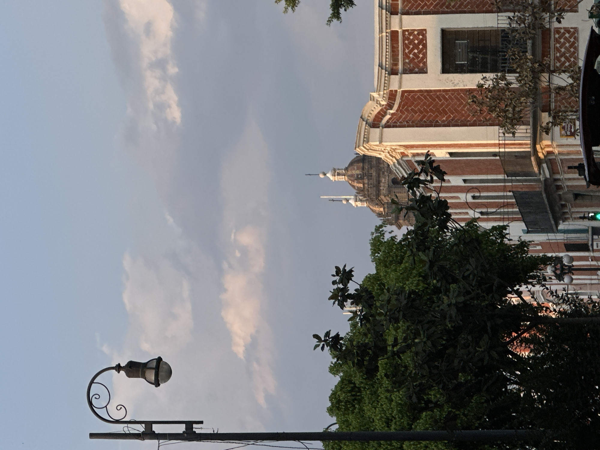 The image shows a building with red brick accents and white trim, potentially a church or religious building, with distinctive domes or spires on top. The sky is visible in the background with scattered clouds. A street lamp with a curved design is visible in the lower left of the frame. Green trees partially obscure the building. The image's location is Unknown, Unknown.
