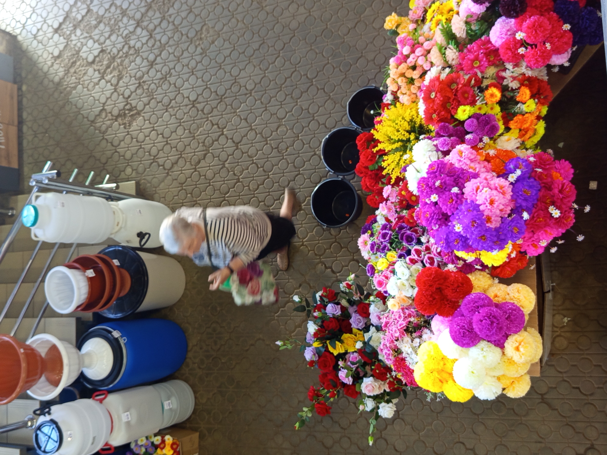 An overhead, eye-level shot captures a bustling outdoor market scene in Gdov, Russia, featuring a woman browsing artificial flowers. The woman, dressed in a striped shirt and dark pants, is seen from above, bending down to inspect a bouquet. She is surrounded by a vibrant display of artificial flowers in a multitude of colors including pink, red, yellow, purple, and white. The flowers are arranged in large bunches and appear to be for sale.
In the background, several large black plastic buckets are visible, some containing more flowers. To the left of the frame, a collection of large white and blue plastic containers, possibly water barrels or storage drums, are stacked and lined up, some with handles and lids. A metal railing with supporting bars runs along the left side of these containers, suggesting they are positioned on or near a staircase or elevated platform. The ground is paved with a light brown, textured surface, possibly tiles or bricks, with a circular pattern.
The image is well-lit, suggesting it was taken during daylight hours, likely in the late morning or afternoon given the general brightness of the scene. No specific weather conditions are evident, but the outdoor setting implies fair weather. The overall mood appears to be one of commercial activity, with the focus on the colorful products and the interaction of the customer. No visible text is discernible in the image.