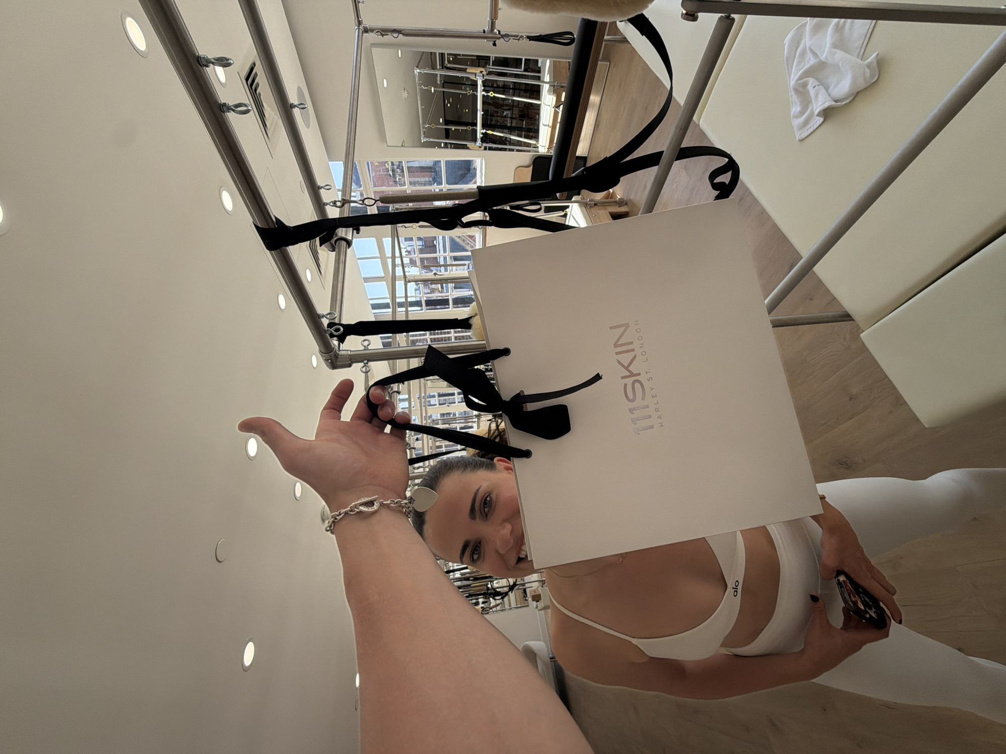 A woman is holding a 111SKIN shopping bag, partially obscuring her face. The bag is white with black handles and the text "111SKIN HARLEY ST. LONDON" printed on it. She is wearing a white sports bra with "ALO" printed on it and white leggings. She is holding a phone in her right hand. She has dark hair pulled back and light skin.
The woman is standing in a pilates studio, with a pilates reformer in the background. The reformer is beige, with a white towel on it. The room has a white ceiling with recessed lighting. Through the windows, buildings and an exterior construction frame can be seen in the city of London.