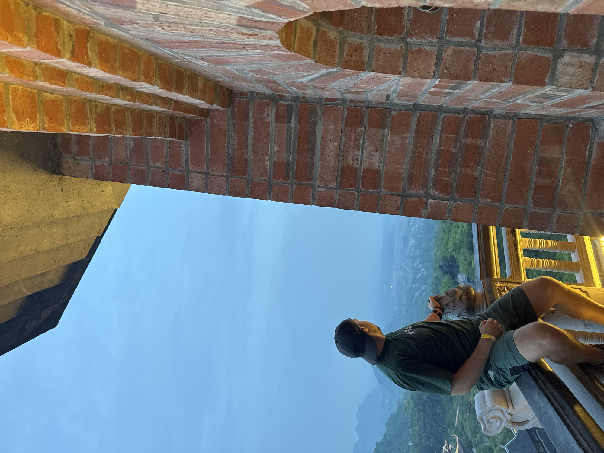 A man wearing a dark green t-shirt, green shorts, and a dark baseball cap is sitting on a railing, looking out at a valley. He is holding a brown bag in his right hand. The railing is made of wood and has decorative white stone elements. Behind him, a brick structure with arched openings is visible. The background shows a hazy landscape of mountains and a town. The sky is overcast and appears to be dusk. The image was taken from a vantage point that overlooks a significant portion of the landscape, likely a temple or a high viewpoint in the Tây Hoa Lư Ward, Vietnam, given the architectural style and the lush mountainous terrain.