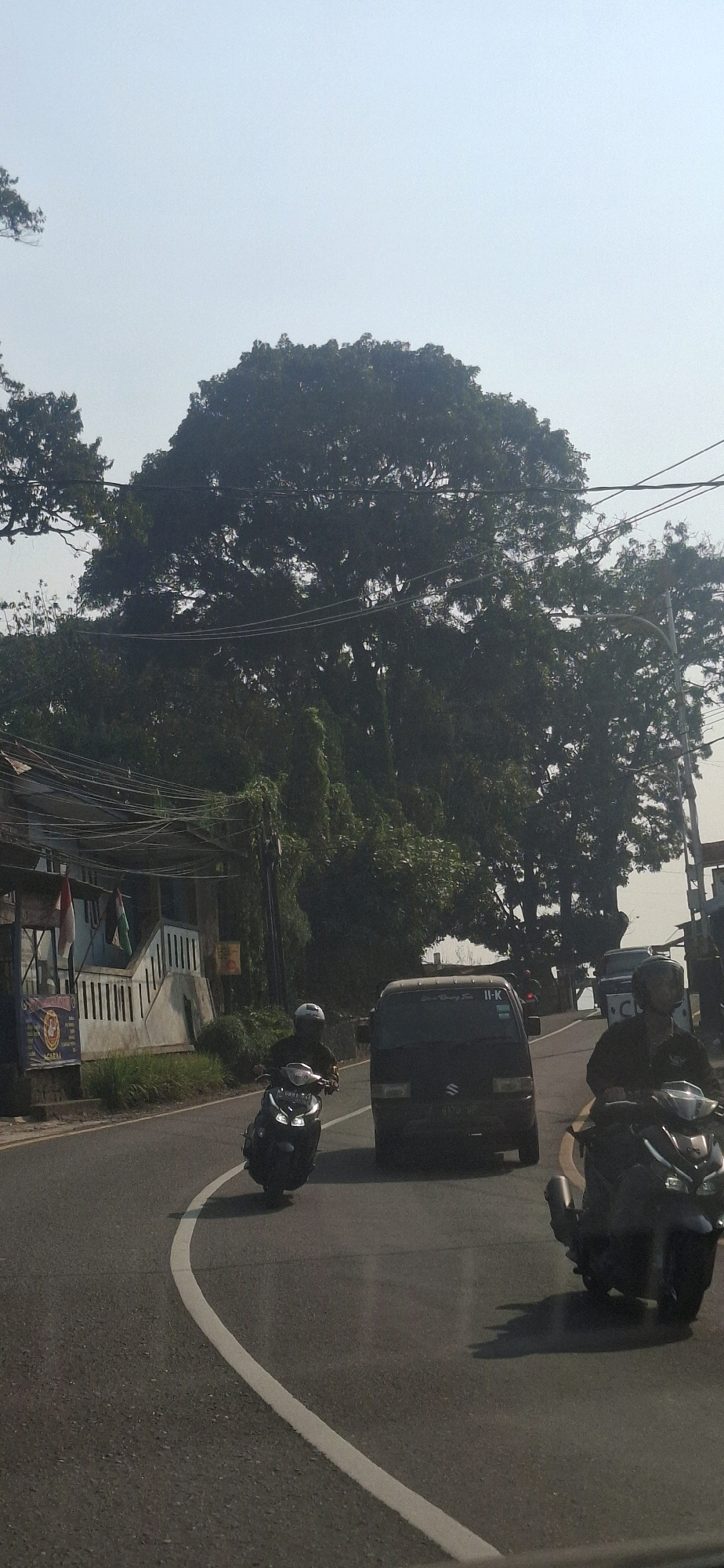 This image depicts a street scene in Cilaku, Indonesia, on a clear day with a hazy sky. A dark-colored van is visible in the center of the frame, driving on a paved road. To the left of the van, a person on a dark scooter with a white helmet is riding in the same direction. On the right side of the road, another person on a dark scooter, also wearing a dark helmet, is in motion, appearing to be behind the van. The road has a white lane marker curving towards the right. On the left side of the road, there is a building with white railings and flags hanging, possibly indicating a residential or commercial establishment. A large, dense tree with dark green foliage dominates the upper half of the image, with power lines running across it. The background consists of more trees and vegetation, suggesting a semi-rural or suburban setting within Cilaku.