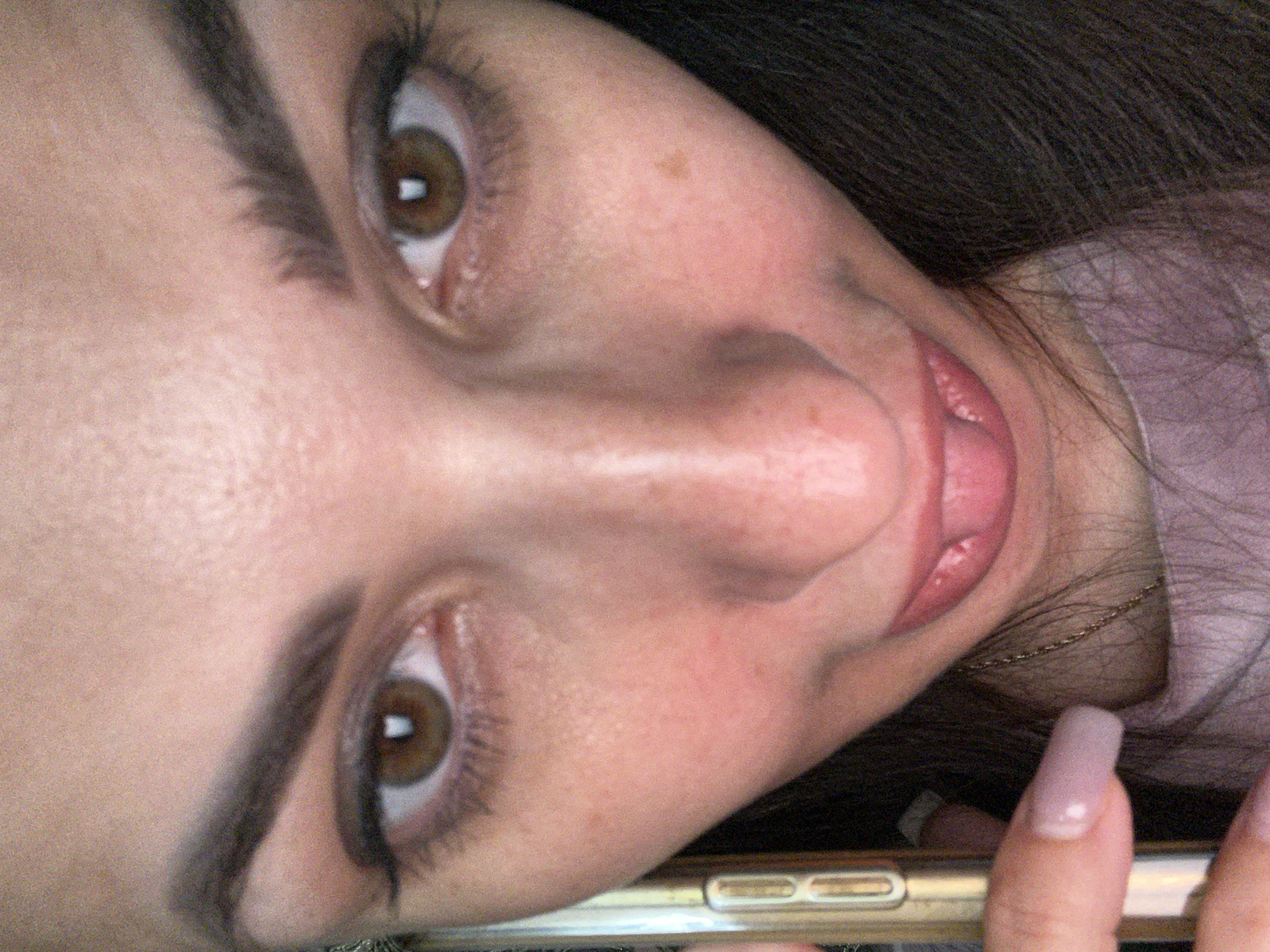 An extreme close-up selfie of a person's face from a low angle, showing their eyes, nose, mouth, and a portion of their forehead and chin. The person has brown eyes, dark eyebrows, and is wearing makeup on their eyes. Their lips are pink and slightly parted, with their tongue visible. A phone is held in the lower left corner of the frame, with a finger with a light pink nail extended towards the viewer. The background is dark, with some of the person's dark hair visible. The image is grainy and has a warm tone.