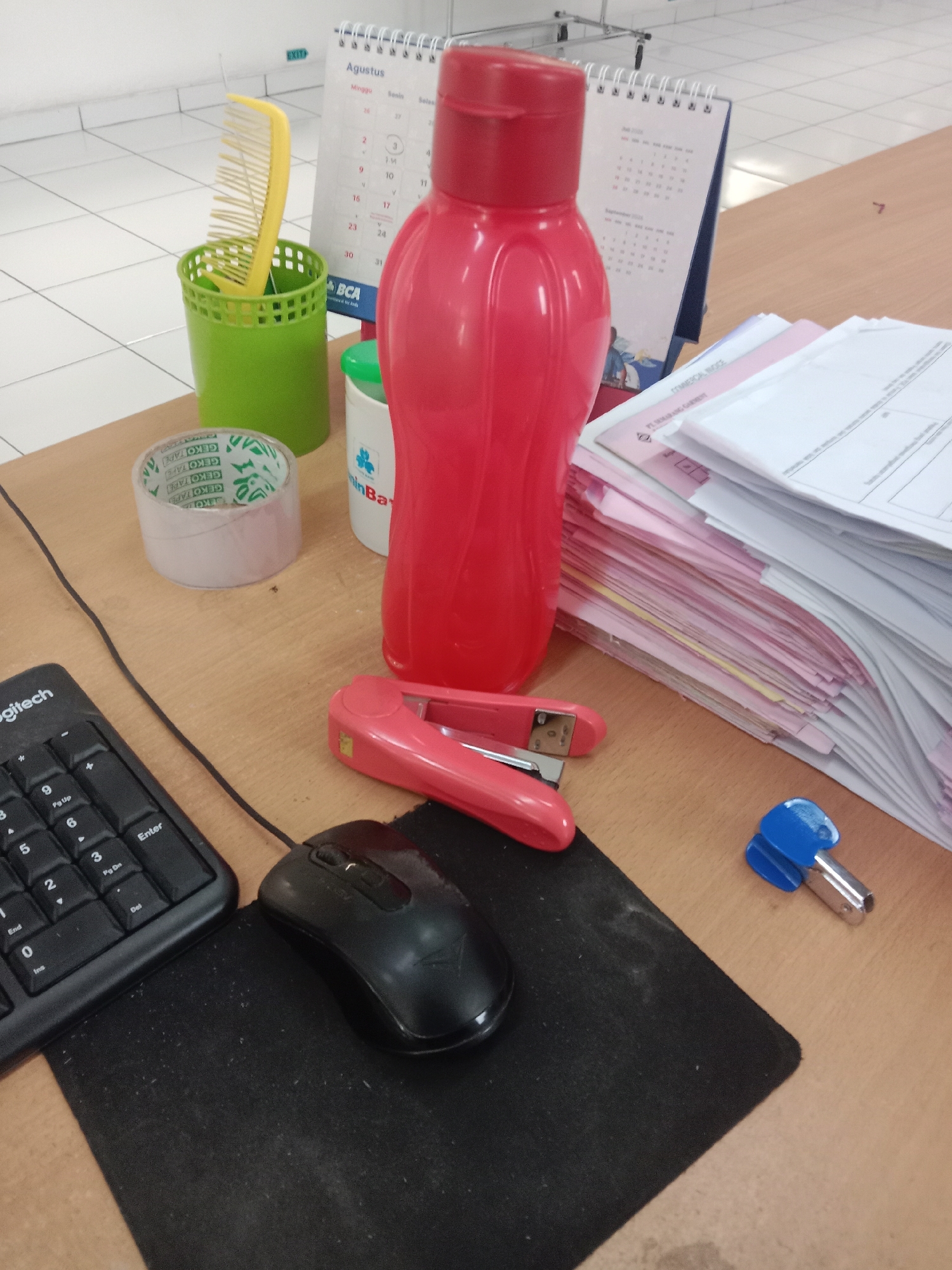 The image depicts a desk in an office environment. On the desk, there is a red water bottle, a yellow comb in a green holder, a roll of tape, a stapler, a keyboard, a mouse on a mousepad, a stack of papers, and a calendar in the background. A blue staple remover is also visible. The calendar displays "Agustus" (August) and shows days of the week in Indonesian. The presence of the Indonesian calendar suggests the location is likely Indonesia. While the specific city of Semarang cannot be definitively confirmed from the image alone, the calendar and its language are consistent with its being a location in Indonesia, and a city like Semarang would be a plausible setting for such an office. There are no people visible in the image, and no actions or interactions are taking place. The environment appears to be a standard office workspace, with tiled walls in the background.