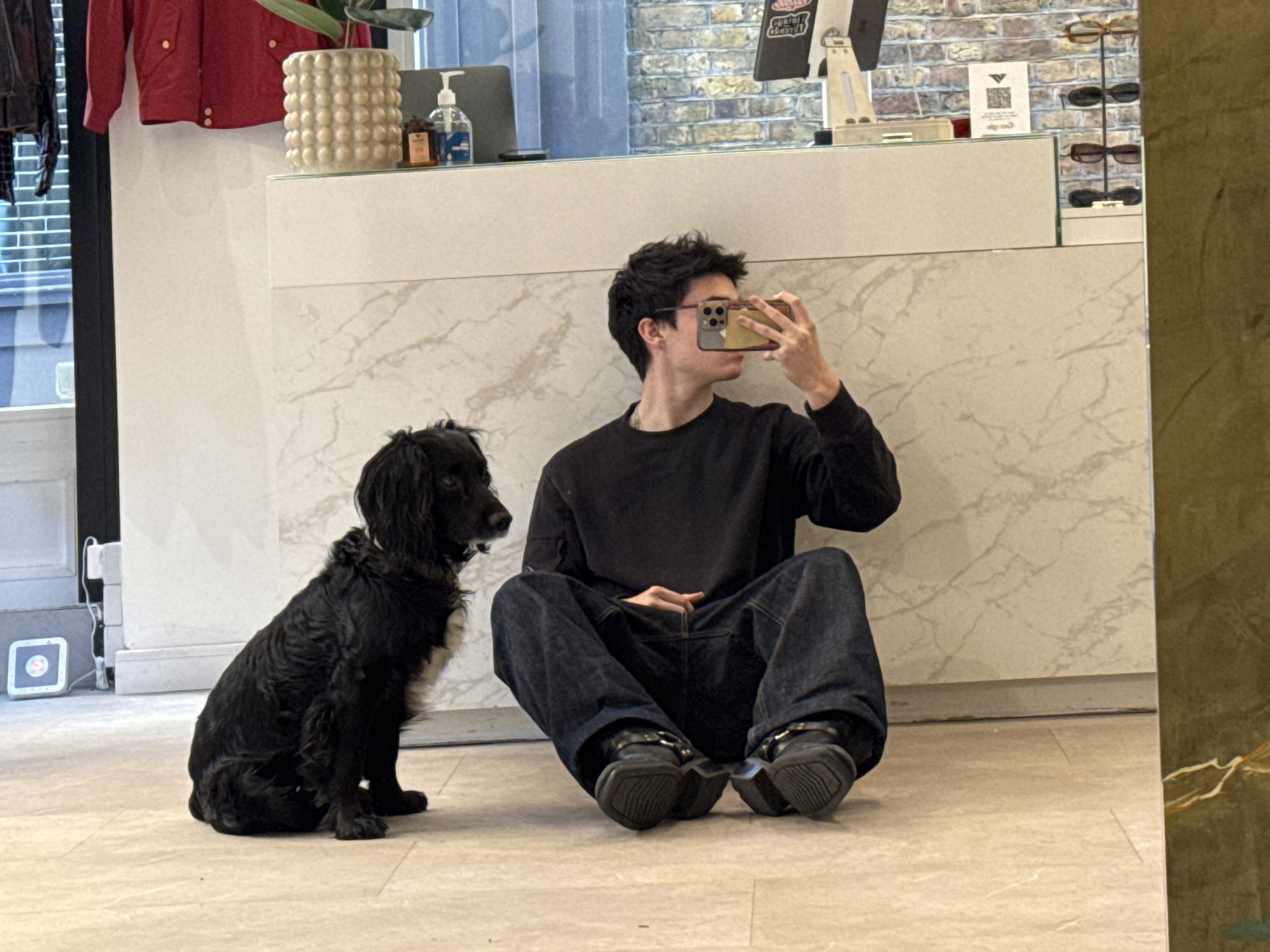 A full shot captures a young man seated cross-legged on a light-colored tiled floor beside a medium-sized black dog. The man is facing slightly to the left, holding a smartphone in front of his face, presumably taking a selfie or photo. He is wearing a long-sleeved black shirt and dark blue jeans, with chunky black shoes. The dog sits to his left, looking directly ahead. Behind them is a white countertop with a marble-like pattern. Visible on the countertop are objects including a plant, hand sanitizer, and what appears to be display stands for sunglasses. In the background, there is a glimpse of a brick wall and an exterior window. The image is taken in a reflective or mirrored space, with a partial view of a wall or surface visible on the right side of the frame. The location is likely a retail space in London, United Kingdom.