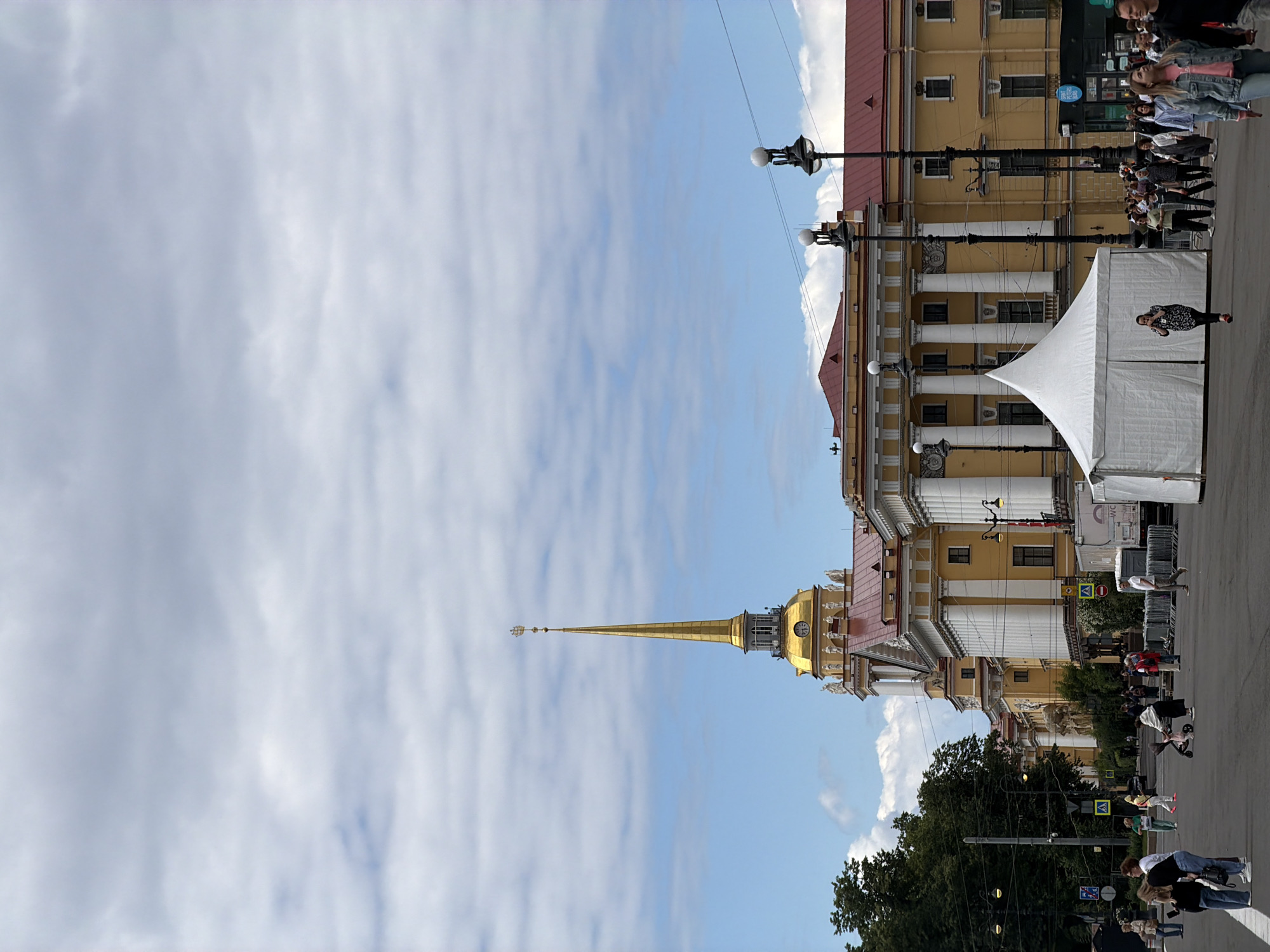 The image depicts an outdoor scene in Saint Petersburg, Russia. The central focus is a golden spire of a building, likely the Admiralty Building, piercing a bright blue sky dotted with wispy clouds. The building itself is large and ornate, painted in shades of yellow and adorned with classical architectural elements such as columns and statues.
In the foreground and along the street, numerous people are visible, appearing to be tourists or locals engaged in various activities. Some are walking, others are standing and looking around, suggesting a public space or a popular landmark. A white tent structure is set up on the street, possibly for an event or market. Lampposts with globes line the street, indicating an urban environment.
The presence of people, the architecture, and the spire strongly suggest a location like Palace Square or a prominent area near the Admiralty. The weather appears to be pleasant, with a clear blue sky and sunlight, indicating it is likely daytime. Some road signs with Russian text are visible, further confirming the location in Russia. The overall mood seems to be one of casual public activity, with people enjoying the architecture and the outdoors.