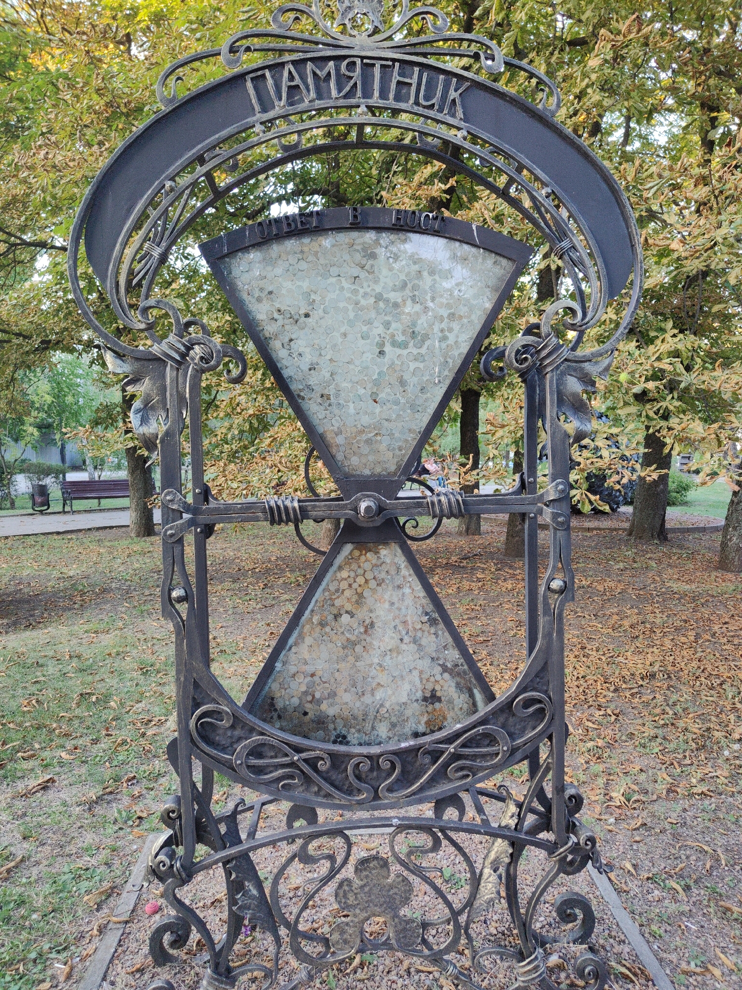 The image shows an outdoor monument in Donetsk, Ukraine. The monument is an hourglass-shaped sculpture made of dark metal with ornate scrollwork. The hourglass chambers are filled with what appears to be coins. A curved metal sign above the hourglass bears Cyrillic lettering that reads "ПАМЯТНИК" (Monument). Below this, another sign reads "В ОТВЕТ В HOC" (In response to HOC), though the full inscription is not entirely clear. The monument is situated in a park-like setting with trees displaying autumn foliage and fallen leaves on the ground. In the background, a park bench and a trash can are visible. The overall scene suggests an artistic installation or memorial within a public space.