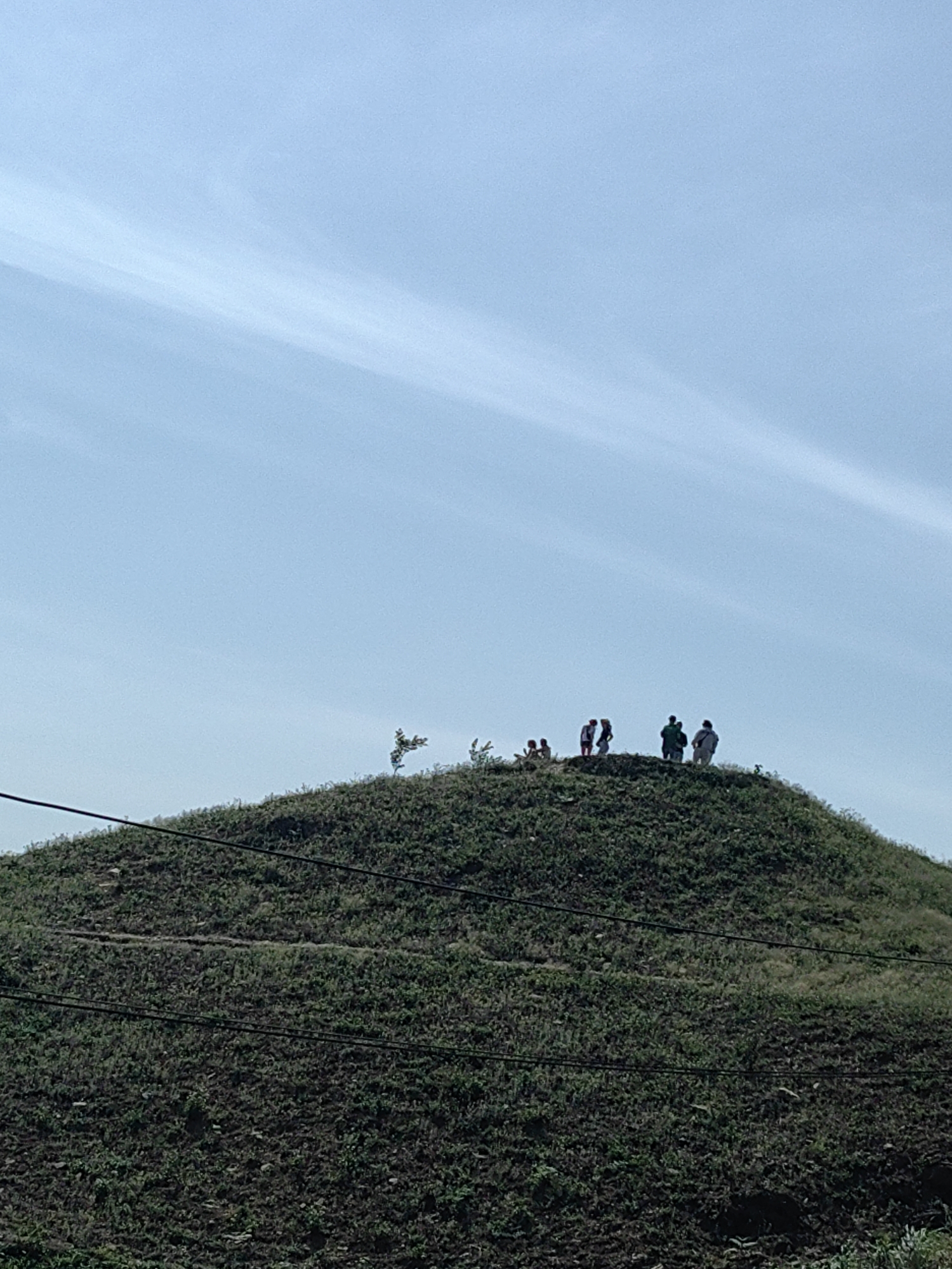 A group of approximately seven people stands atop a grassy hill in Lũng Cú Commune, Vietnam. The sky is pale blue with wispy white clouds. Power lines cross the hill's slope. The hill is covered in vegetation. The ground at the bottom of the hill is dark and appears to be freshly cultivated or disturbed soil. The people are silhouetted against the sky.