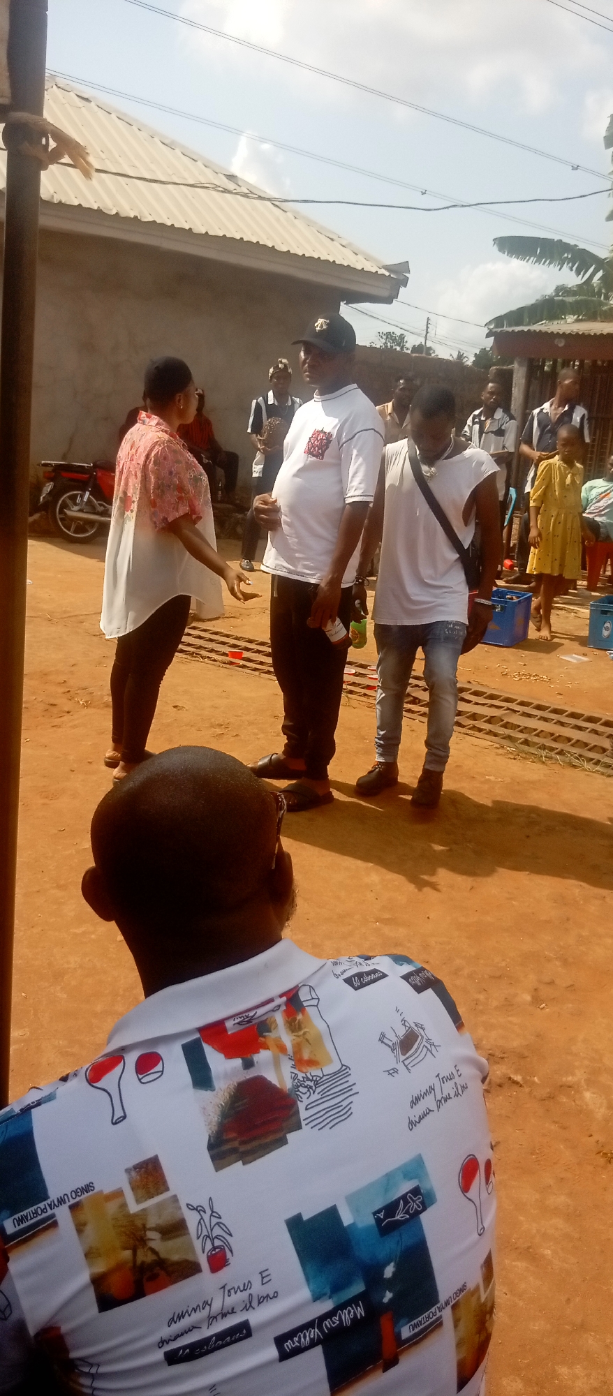 The image depicts an outdoor scene in Ogidi, Nigeria, featuring several people in a casual gathering. A man wearing a white T-shirt and black cap stands talking to a woman in a white and pink floral-patterned shirt. Another man in a white tank top and jeans stands nearby. In the foreground, the back of a seated person wearing a white patterned shirt is visible. The setting appears to be a dusty or dirt-covered area, with buildings, a motorcycle, and blue crates visible in the background, indicating an outdoor event or meeting. Other people are standing or sitting in the background, suggesting a group setting.