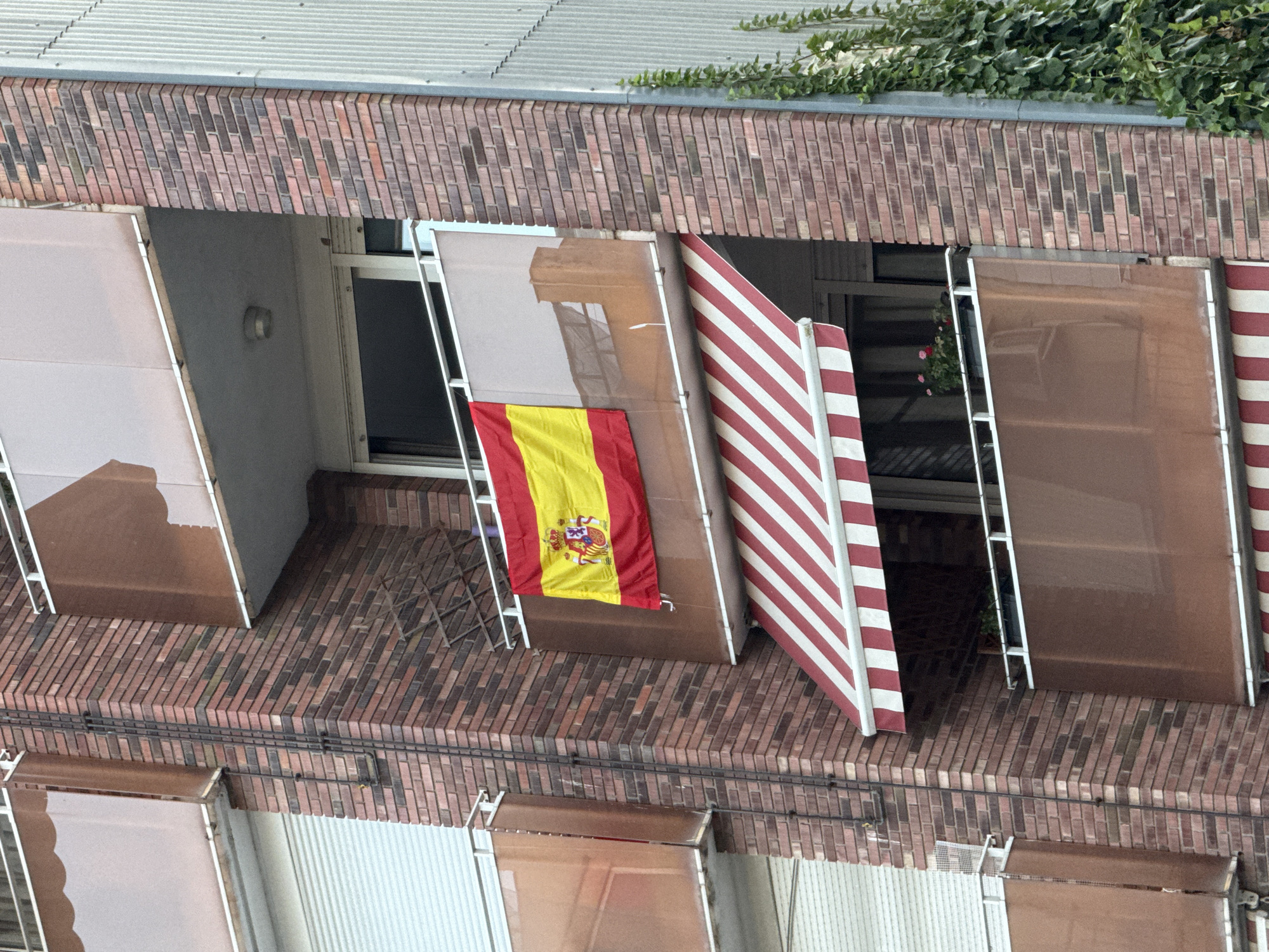 The image shows the facade of a brick apartment building in Blanes, Spain. A Spanish flag is displayed on a balcony, hanging from a railing. The flag is predominantly yellow and red, with the coat of arms of Spain visible in the center. Below this balcony, there is a striped awning, red and white, which extends outward. The building has multiple balconies with translucent brown panels. The overall environment appears to be a residential area.