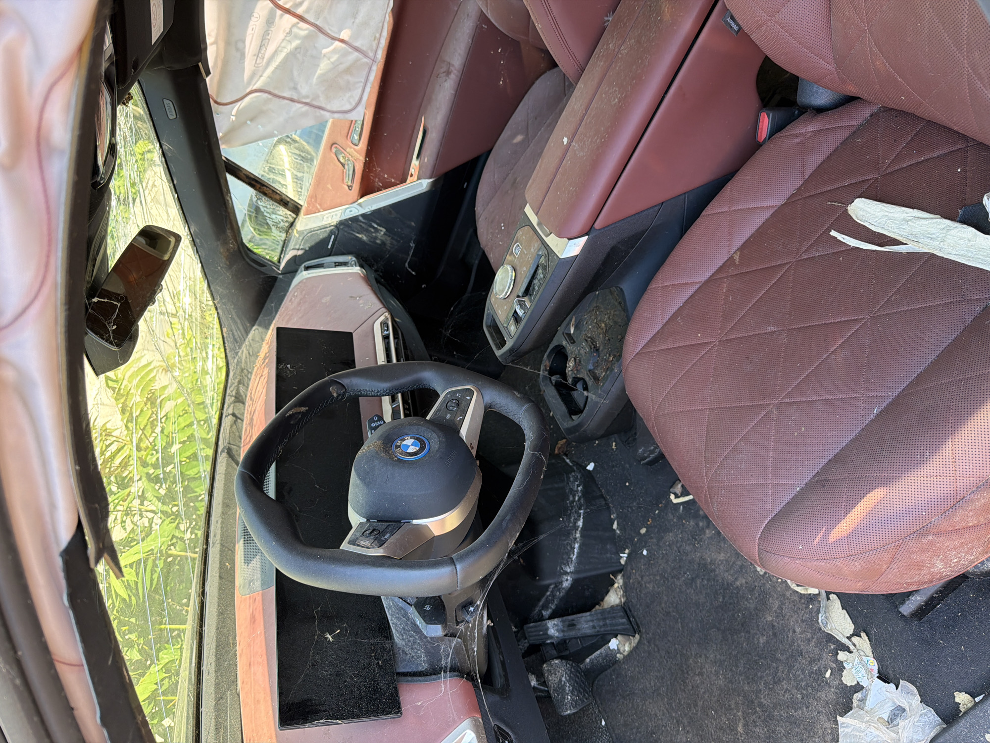 The image shows the interior of a severely damaged luxury car, likely a BMW iX, parked outdoors. The windshield is cracked, and the airbags have deployed, indicating a recent accident. Debris is scattered throughout the interior, including on the seats and floor. The car's interior is a deep burgundy color, with a large digital display on the dashboard. The exterior is visible through the broken windshield, showing greenery and what appears to be a bright sky, suggesting a sunny day. The scene is consistent with a car that has been abandoned or is awaiting salvage after a significant collision, and it is positioned in an urban or semi-urban environment, possibly a junkyard or impound lot. No people are visible in the image.
