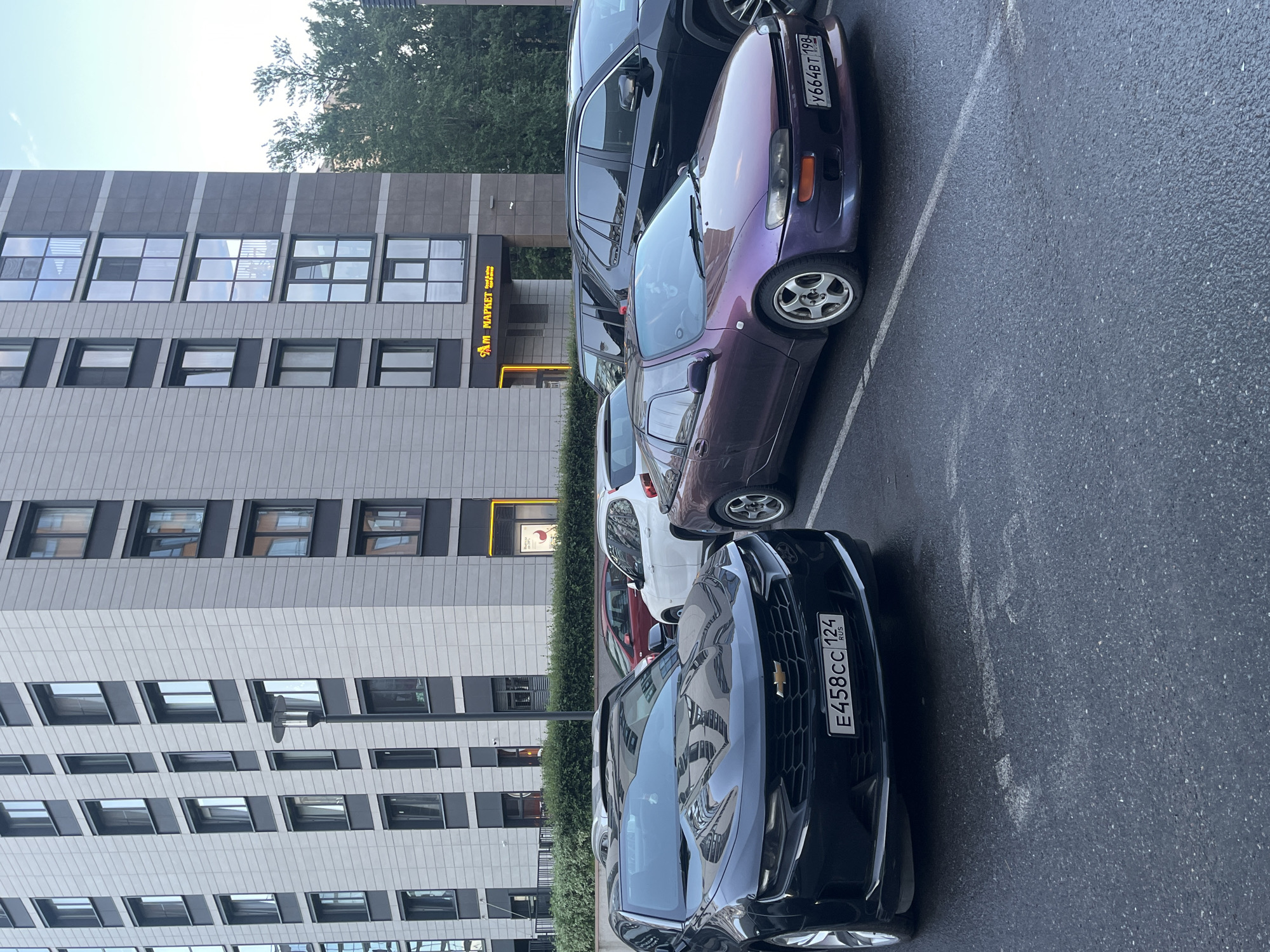 The image depicts an outdoor parking lot in front of a modern, multi-story residential or commercial building. The building has a facade of grey tiles and dark, recessed window frames. A prominent sign with yellow lettering reads "AM MAPKET". Several cars are parked in the lot, including a black Chevrolet Camaro in the foreground with a Russian license plate "E458CC 124". To the right of the Camaro is a purple sports car with a Russian license plate "Y664BT 198". Behind these cars are other vehicles, including a white hatchback and a dark SUV. The pavement is wet, suggesting recent rain or dew. Green foliage is visible behind the building on the right side of the image.
