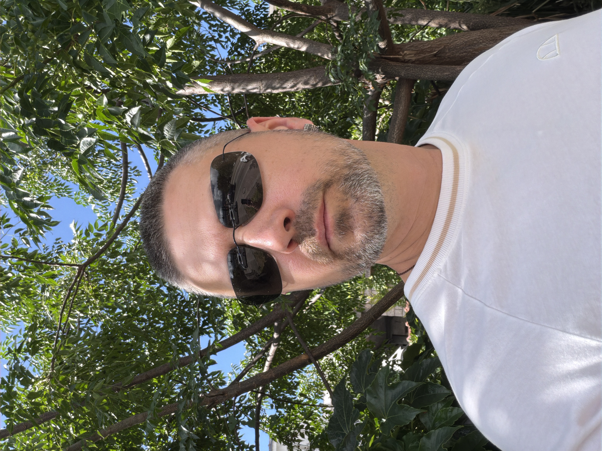 A portrait-style image captures an adult man, likely in his 30s or 40s, posing outdoors under a dense canopy of green trees in Budva, Montenegro. The man's head is tilted, and he looks directly at the camera, his face dominating the left side of the frame. He has short, dark hair with visible grey stubble or a short beard. Dark sunglasses cover his eyes, reflecting parts of the bright, sunny surroundings. A small ring or earring adorns his left ear. He is wearing a white, short-sleeved t-shirt featuring light brown or gold horizontal stripes on the collar/cuff, and a subtle white embroidered logo, possibly an abstract 'A' or shield shape, on the chest. The background consists of vibrant green foliage and glimpses of a clear blue sky, suggesting a bright midday or early afternoon setting with pleasant weather. The overall impression is one of casualness or leisure.