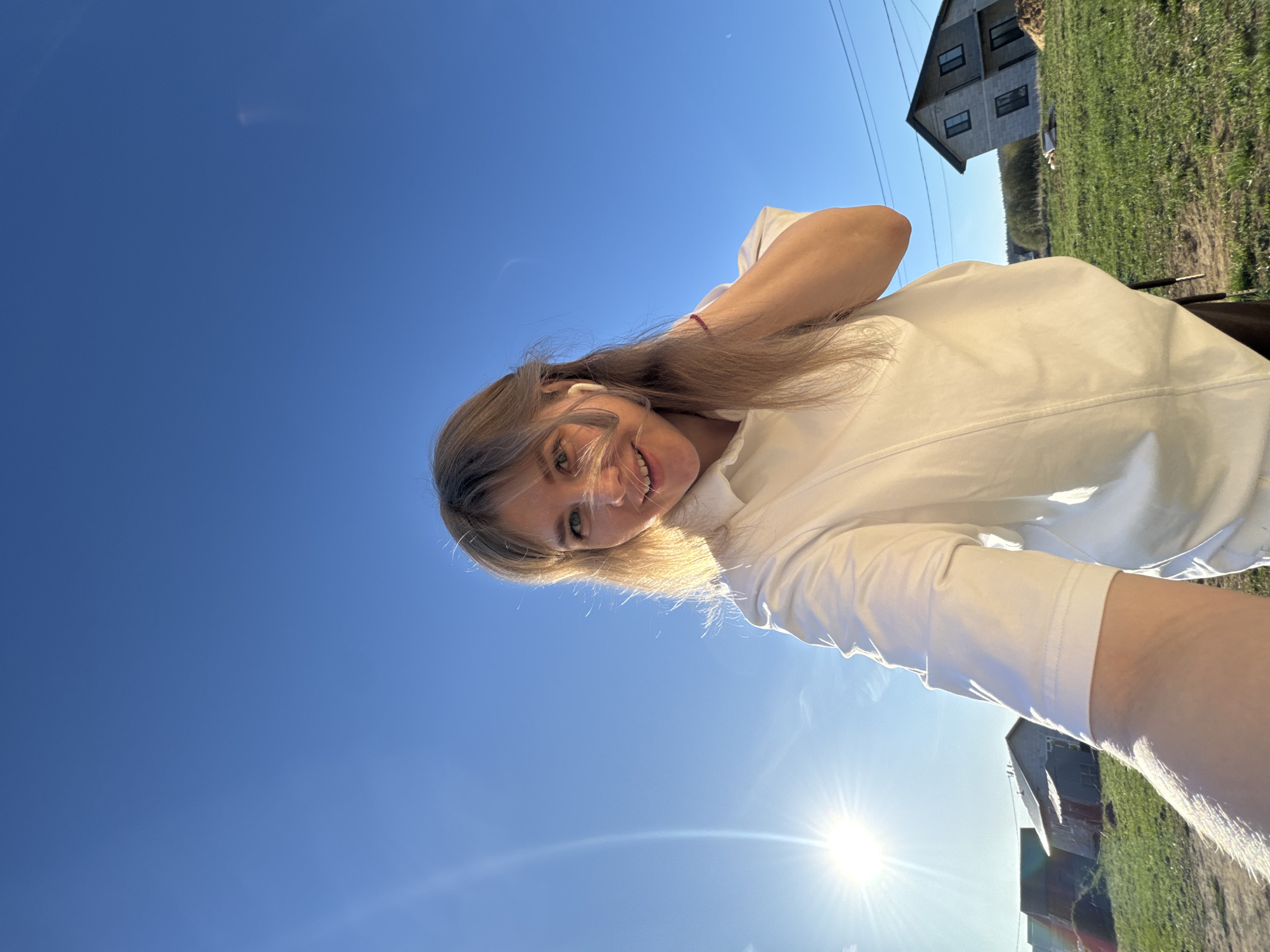 A low-angle shot shows a young woman with blonde hair smiling towards the camera. She is wearing a white shirt and has what appears to be a wireless earbud in her ear. The background features a bright blue sky with the sun shining. In the distance, a house with grey siding and a green lawn can be seen. The location is likely the city of Ферма, Russia, based on the prompt's context. The sun is positioned near the bottom of the frame, creating a lens flare effect.
