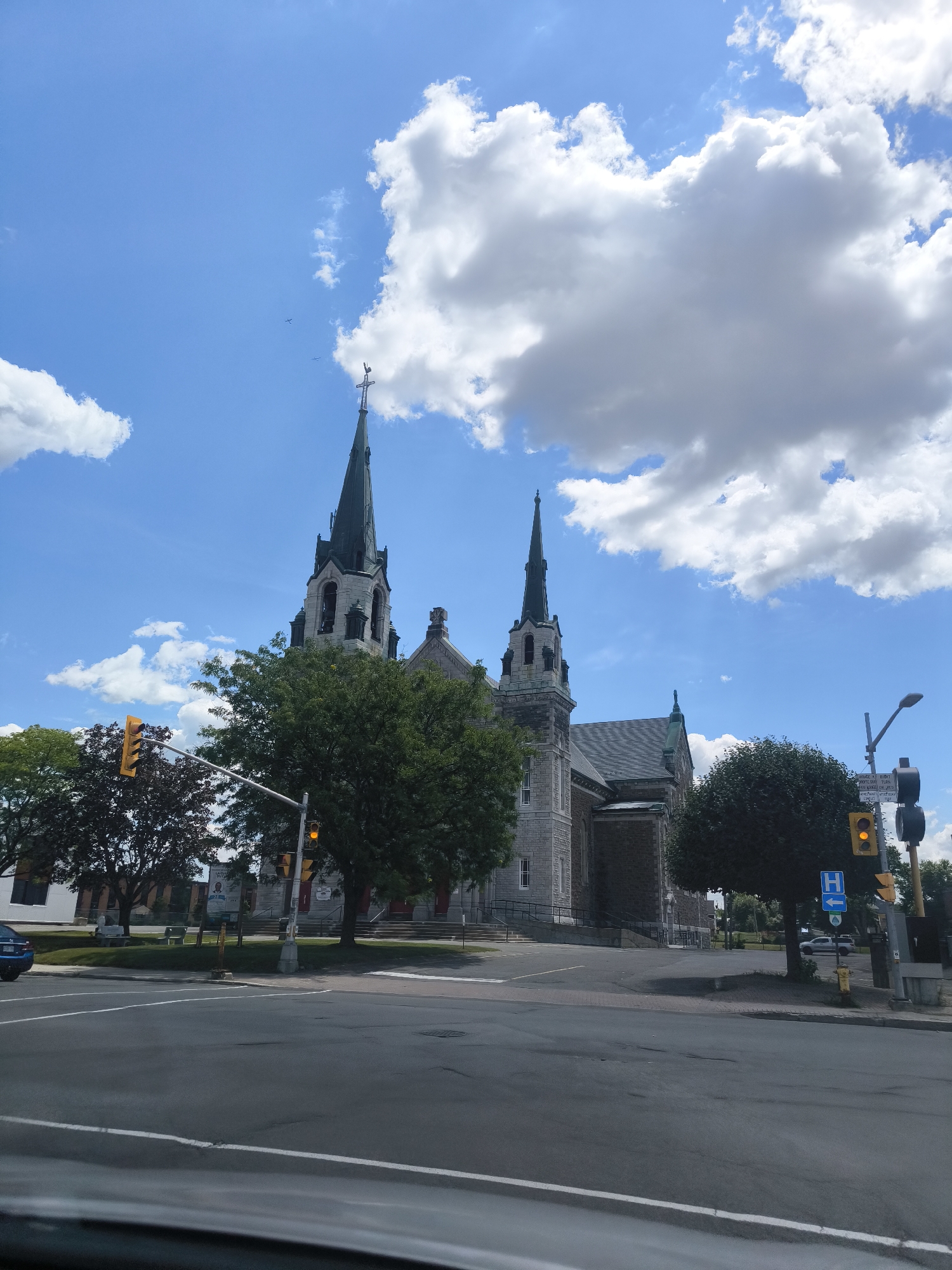 This is a wide-angle, eye-level outdoor shot taken on a clear, partly cloudy day. The main subject is the stone facade of a church with two prominent steeples, one taller and more pointed than the other. The steeples are dark green, and the church itself is constructed of light-colored stone with darker accents and a dark gray roof. A green cross sits atop the taller steeple. Lush green trees partially obscure the view of the church, with one particularly dense tree in the foreground on the right.
In the foreground, a street intersection with asphalt paving and white lane markings is visible. Traffic lights are present, with one displaying a yellow light. A blue sign with a white "H" indicates a hospital. A white line marking the edge of the road runs parallel to the bottom of the frame.
The environment is urban, with other buildings visible in the background, though less detailed. The sky is a vibrant blue, dotted with large, fluffy white clouds. The scene appears to be captured from within a vehicle, given the slight blur at the bottom edge, suggesting a road or dash view.
This church is located at the intersection of John Street and Main Street in Hawkesbury, Ontario, Canada.