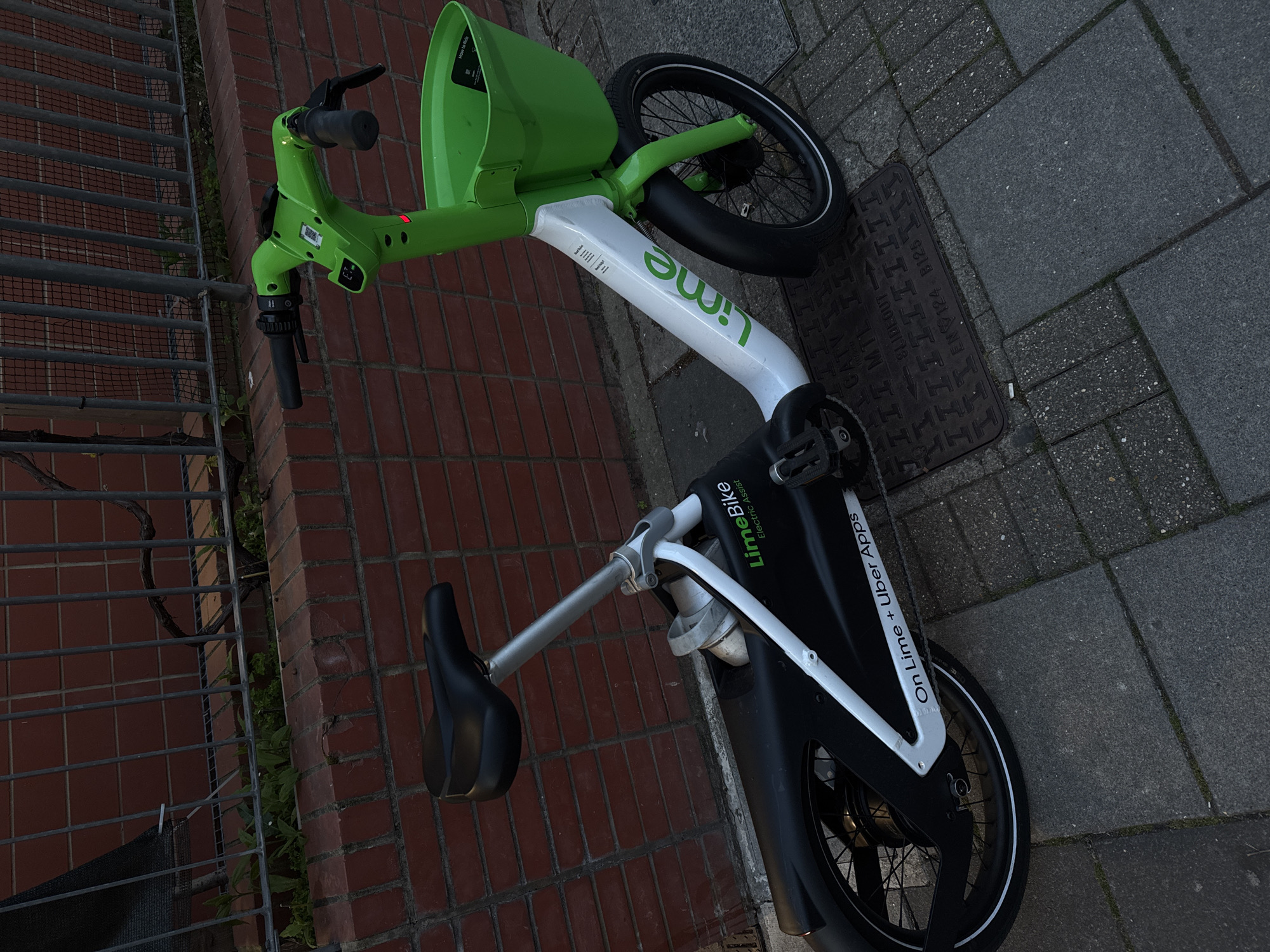 A bright green and white Lime electric assist bicycle is depicted lying on its side on a paved sidewalk, rotated 90 degrees clockwise in the image. The bike features a prominent green front basket and handlebars, a white main frame with "Lime" written in green, and a black rear section. The setting is an urban environment, likely in London, United Kingdom, with a dark red brick wall and a grey metal fence running along one side of the pavement. The ground consists of grey concrete paving slabs and a rectangular metal manhole cover.
The lighting indicates daytime, possibly overcast or late afternoon, with soft, diffused shadows. The bike appears stationary, having been left or fallen over. No individuals are visible.
Visible text on the bike includes "LimeBike," "Electric Assist," and "On Lime + Uber Apps" on the black section of the frame, and "Lime" displayed prominently on the white frame. The basket also shows partial text, "How to Ride." The manhole cover is inscribed with "SLIDEOUT-11," "CATV MTL," and "EN 124 B125," along with recurring patterns of vertical lines.